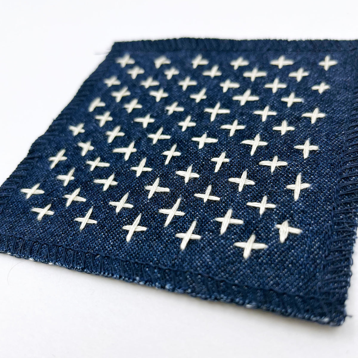 A square denim patch of with and embroidered rows of ivory plus signs, displayed on a clean white background.