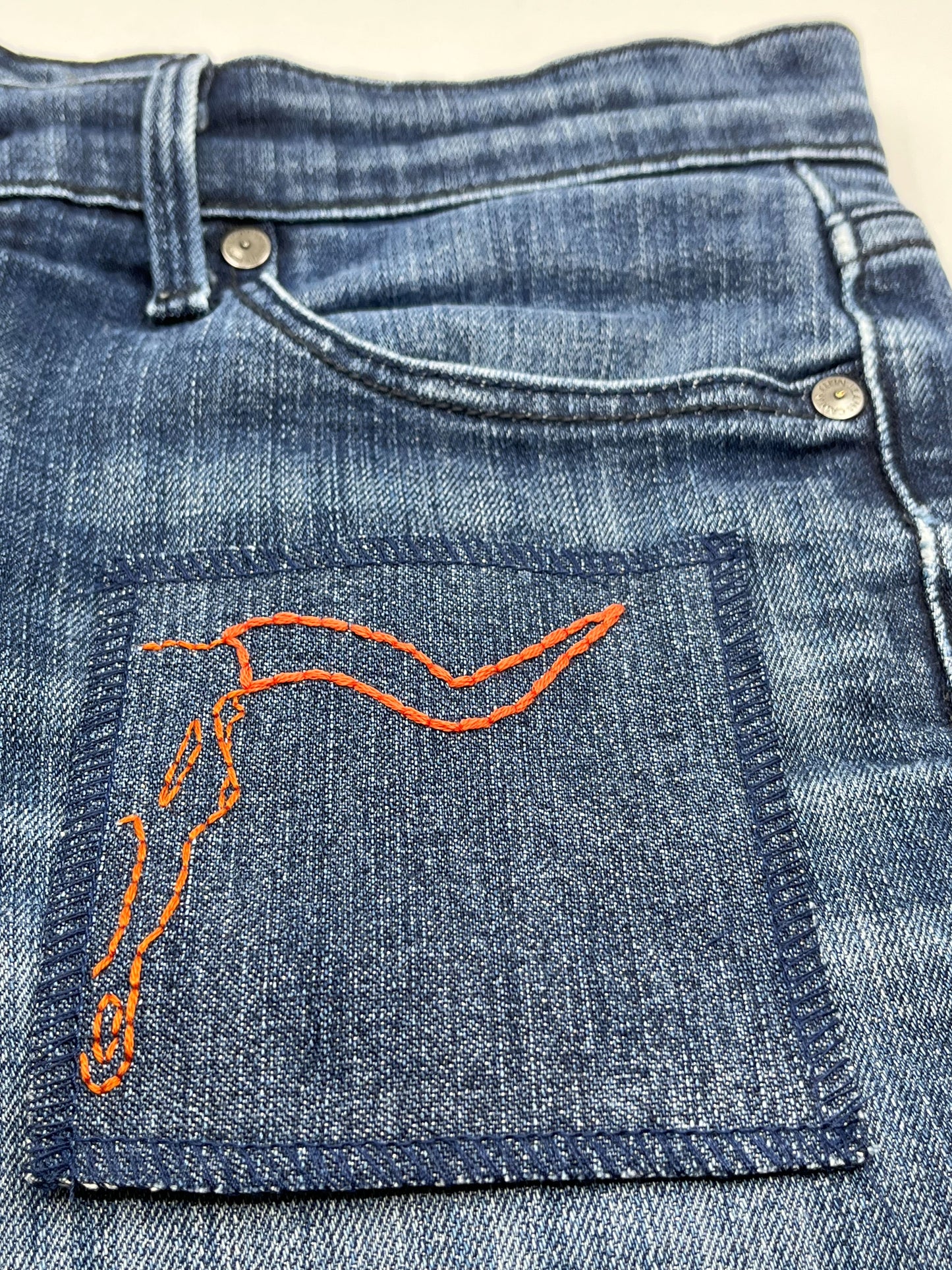 a square denim patch with overlocked edges, with the right side of a longhorn skull hand embroidered on it, in rust colored thread with backstitch and french knots, on a pair of jeans