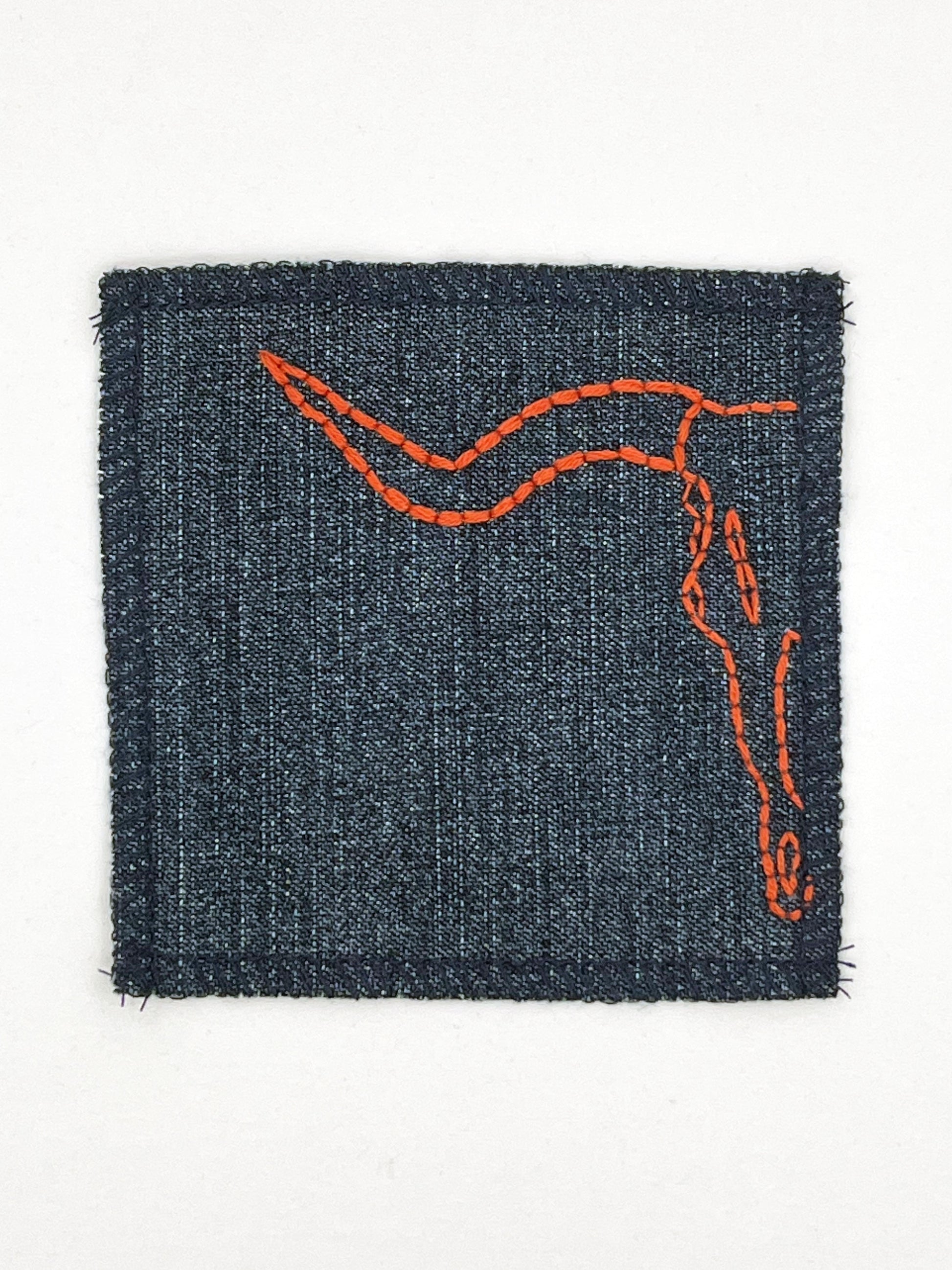 close up view of a square denim patch with overlocked edges, with the left side of a longhorn skull hand embroidered on it. in rust thread with backstitch and french knots, on a white background