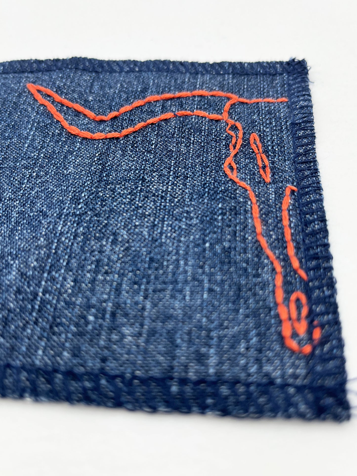 close up view of a square denim patch with overlocked edges, with the left side of a longhorn skull hand embroidered on it. in rust thread with backstitch and french knots, on a white background
