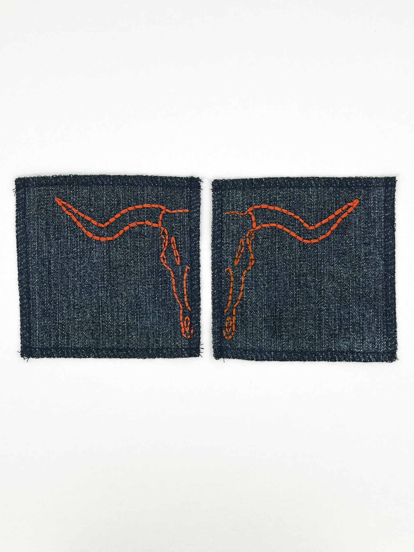 two square denim patches with overlocked edges, with longhorn skulls hand embroidered on them, one patch has the left side of the skull and the other has the right side. in rust thread with backstitch and french knots, on a white background