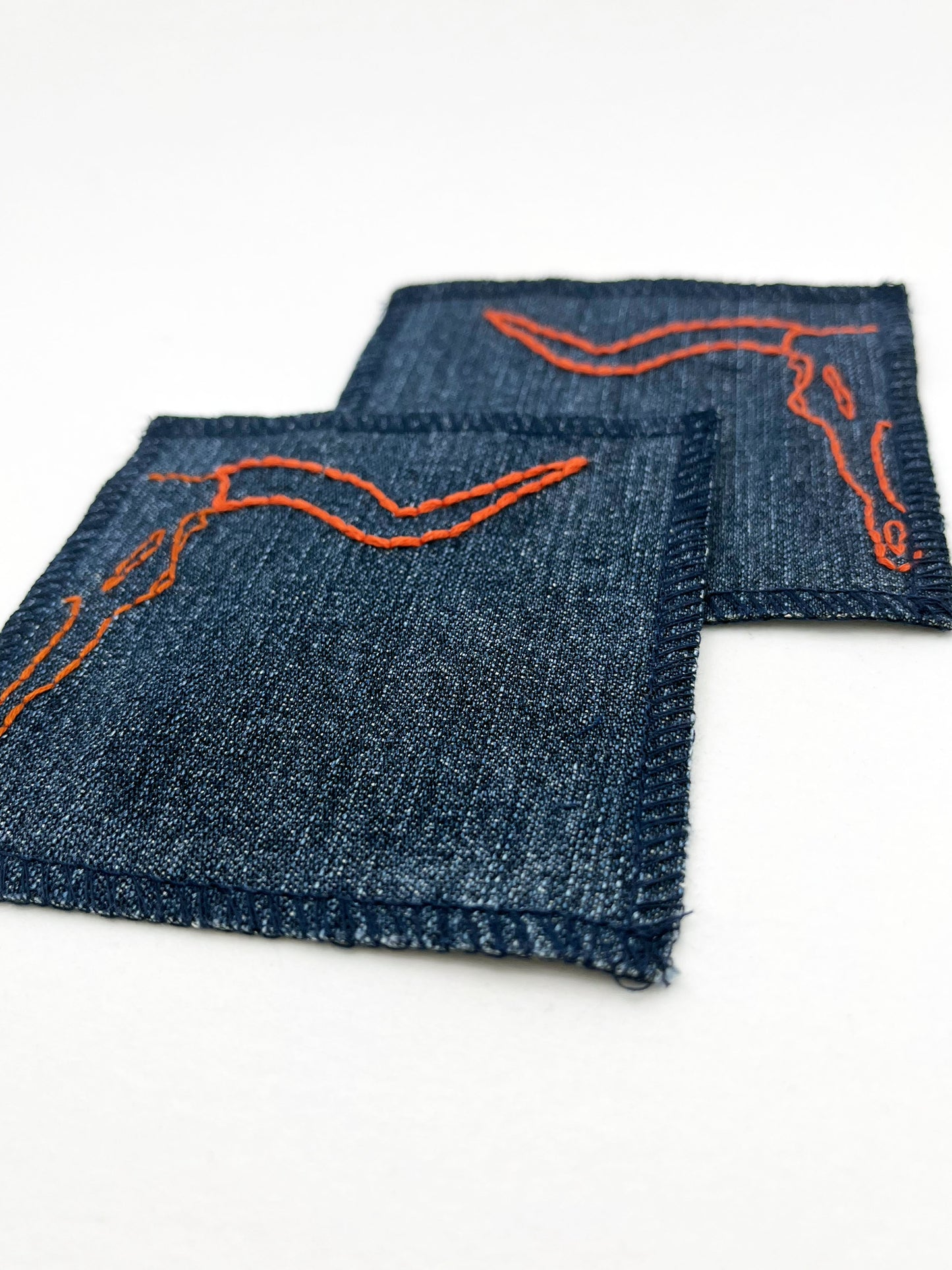 two square denim patches with overlocked edges, with longhorn skulls hand embroidered on them, one patch has the left side of the skull and the other has the right side. in rust thread with backstitch and french knots, on a white background