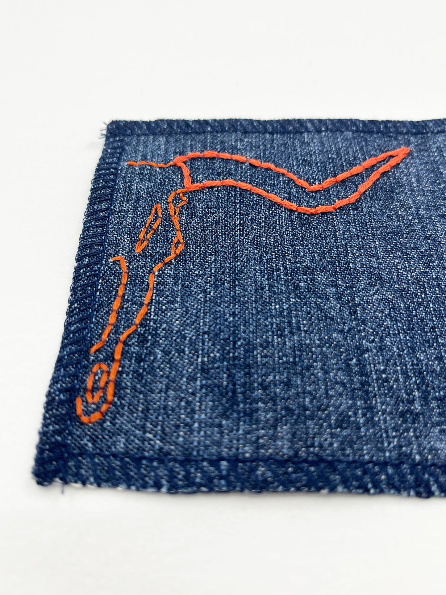 close up view of a square denim patch with overlocked edges, with the right side of a longhorn skull hand embroidered on it. in rust thread with backstitch and french knots, on a white background