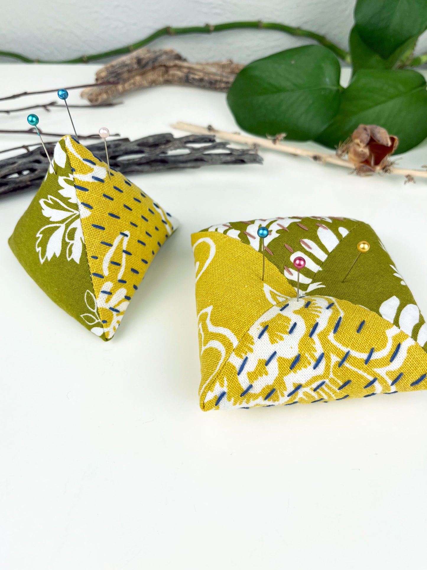 two pincushions one a pyramid shape and the other a square pillow made from floral olive and mustard fabric with contrasting sashiko stitches on a white counter with dried twigs seedpods and a pothos in the background