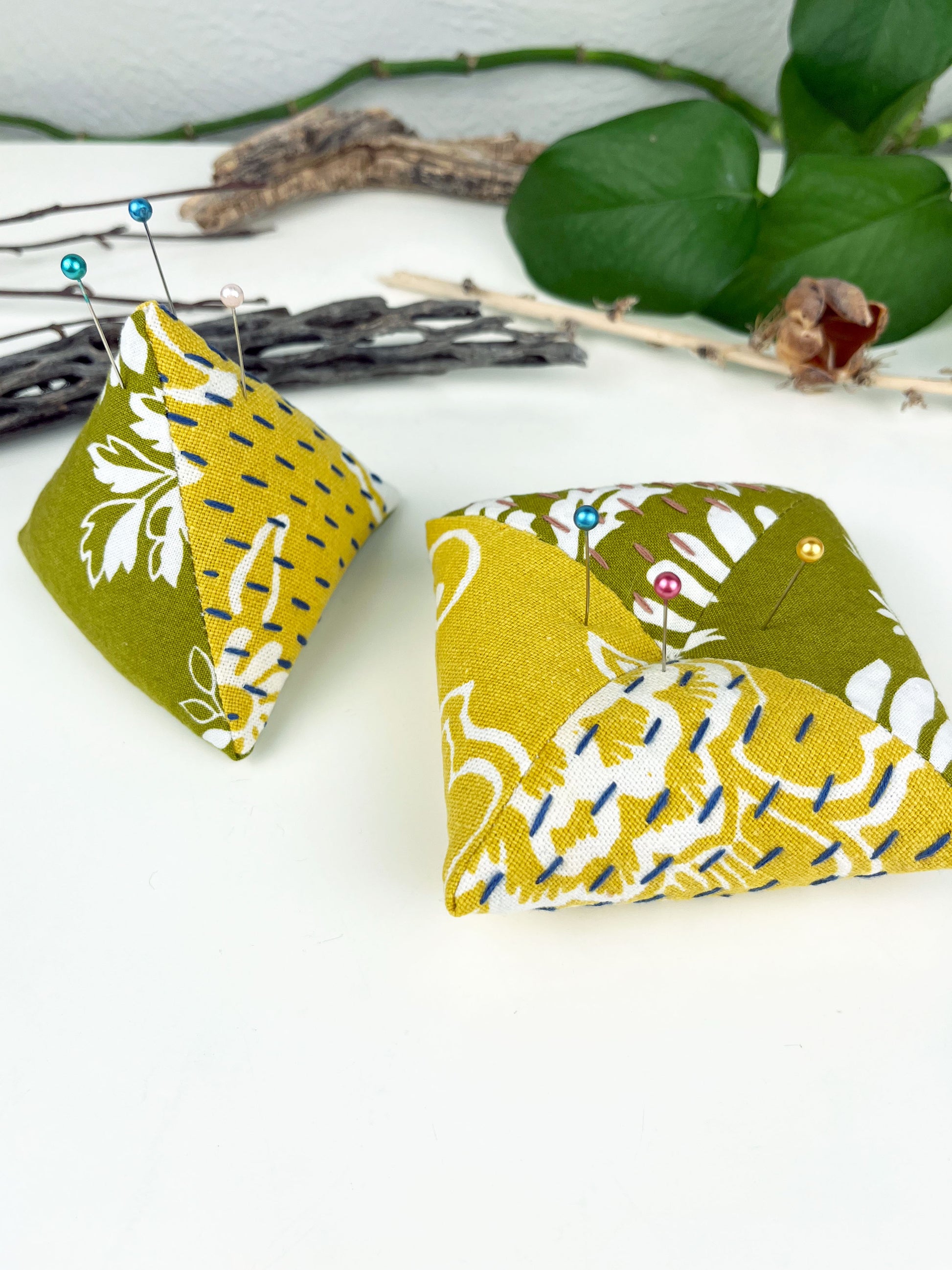 two pincushions one a pyramid shape and the other a square pillow made from floral olive and mustard fabric with contrasting sashiko stitches on a white counter with dried twigs seedpods and a pothos in the background