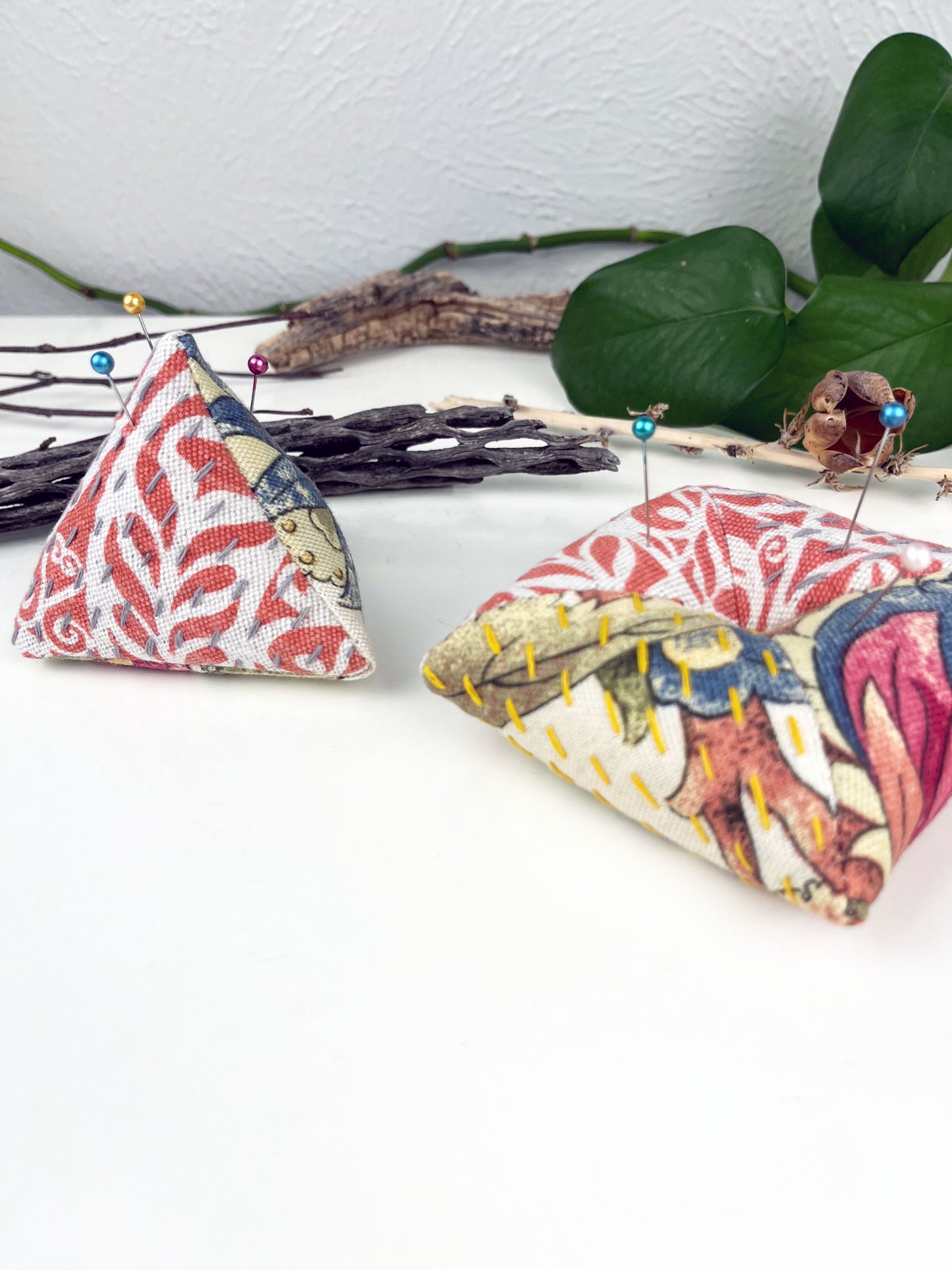 two pincushions one a pyramid shape and the other a square pillow made from floral and orange upholstery fabric with contrasting sashiko stitches on a white counter with dried twigs seedpods and a pothos in the background