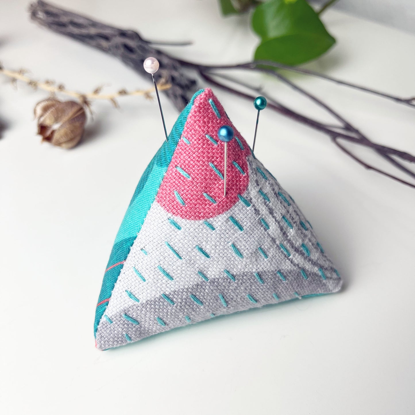 a pyramid pincushion made from aqua fuchsia and gray patterned fabric with contrasting sashiko stitches on a white counter with dried twigs seedpods and a pothos plant in the background