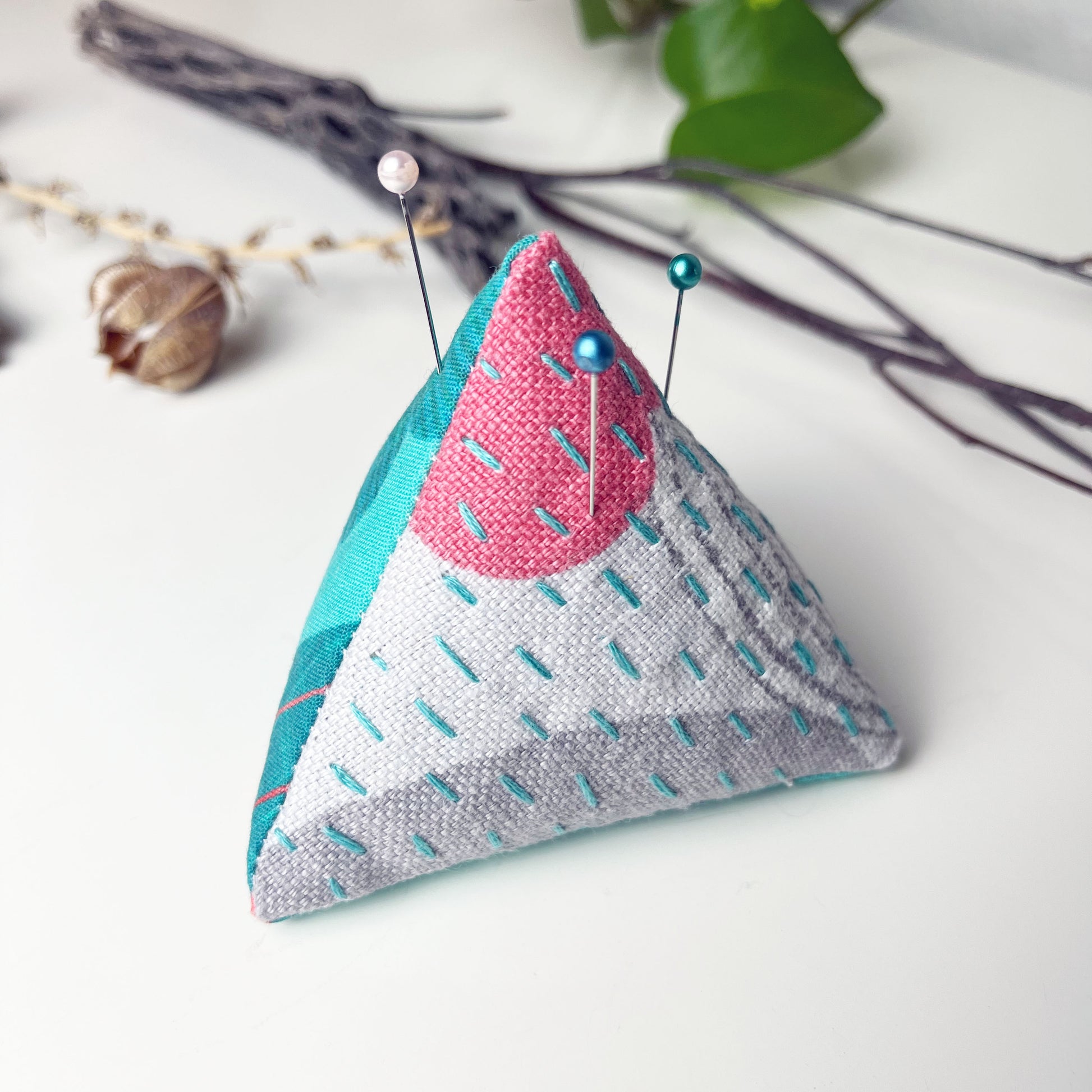 a pyramid pincushion made from aqua fuchsia and gray patterned fabric with contrasting sashiko stitches on a white counter with dried twigs seedpods and a pothos plant in the background