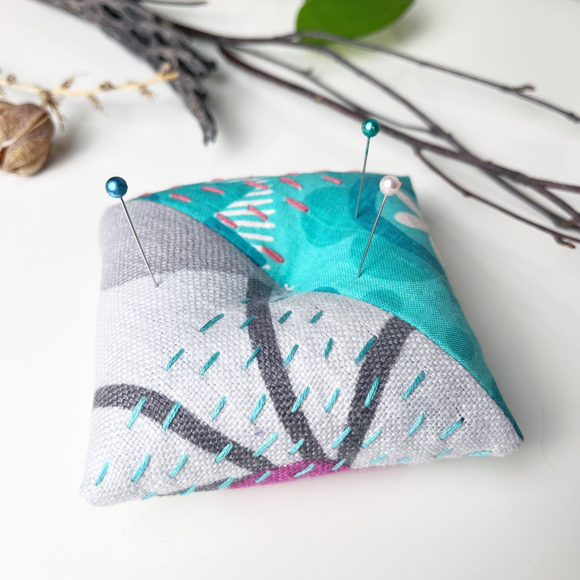 a square pillow shaped pincushion made from aqua gray and fuchsia patterned fabric, with sashiko stitching in contrasting colors, on a white counter with dried twigs seedpods and a pothos plant in the background