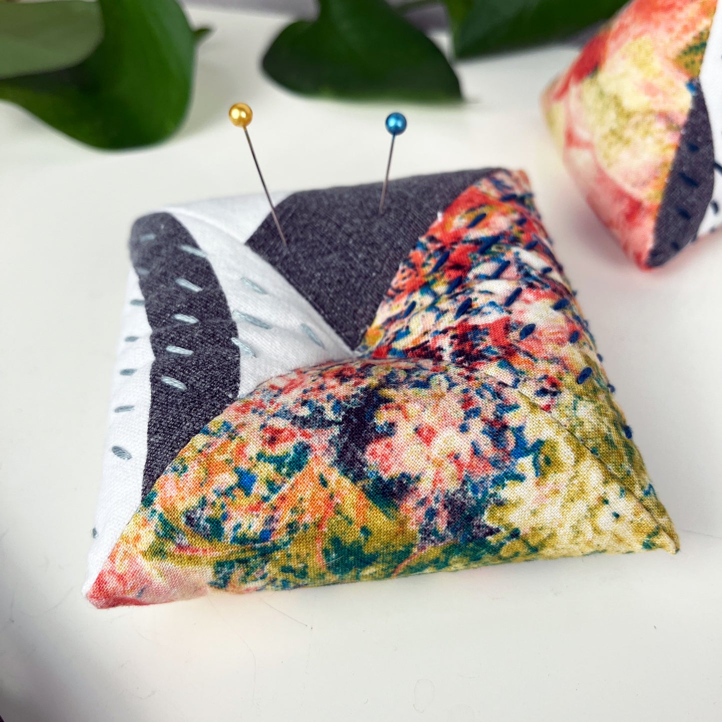 a square pincushion made from multi colored and black and white fabric with contrasting sashiko stitches on a white counter 