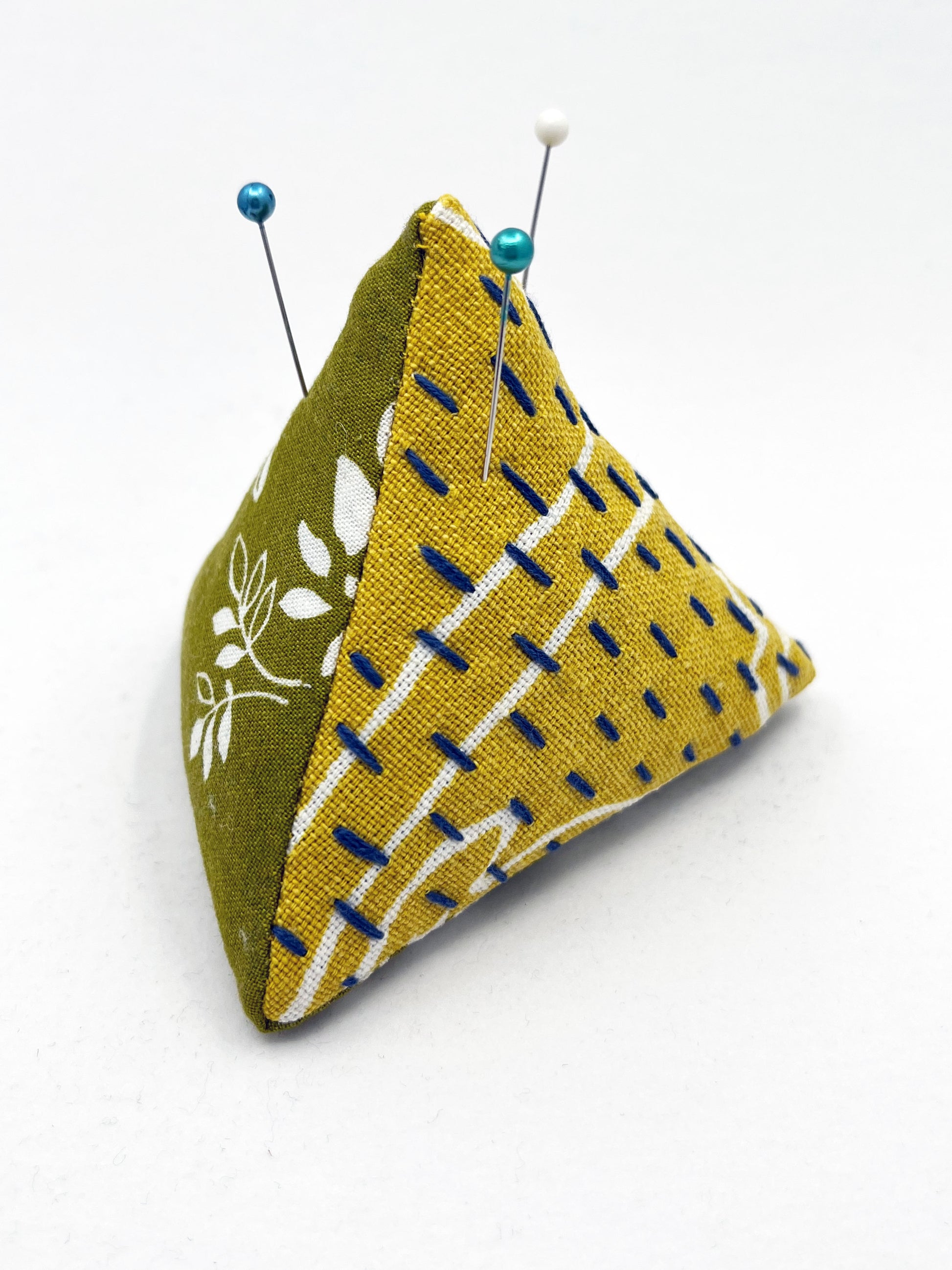 a pyramid pincushion made from floral olive and mustard colored fabric with contrasting sashiko stitches on a white counter 