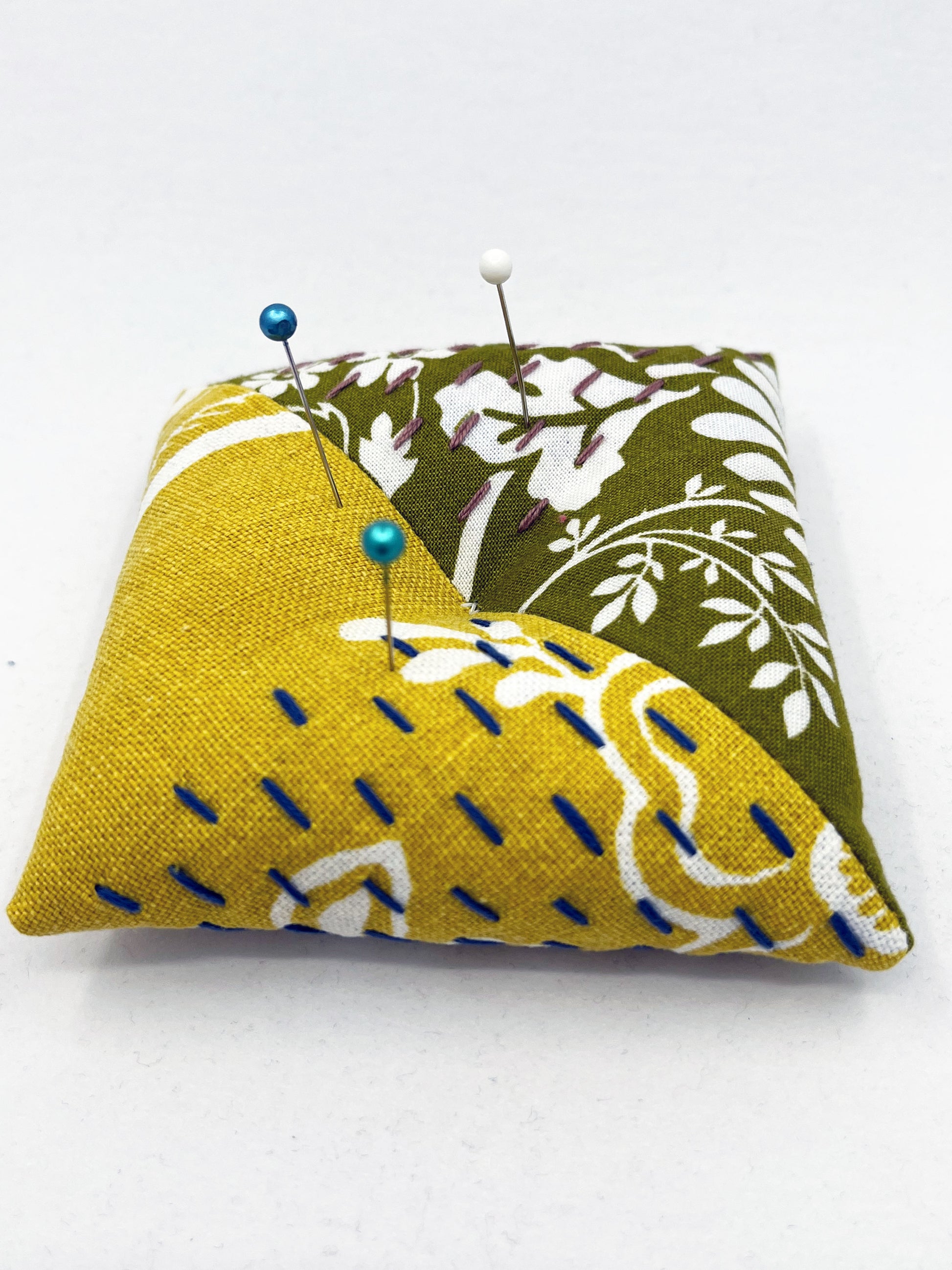 a square pillow shaped pincushion made from floral olive and mustard colored fabric with contrasting sashiko stitches on a white counter 