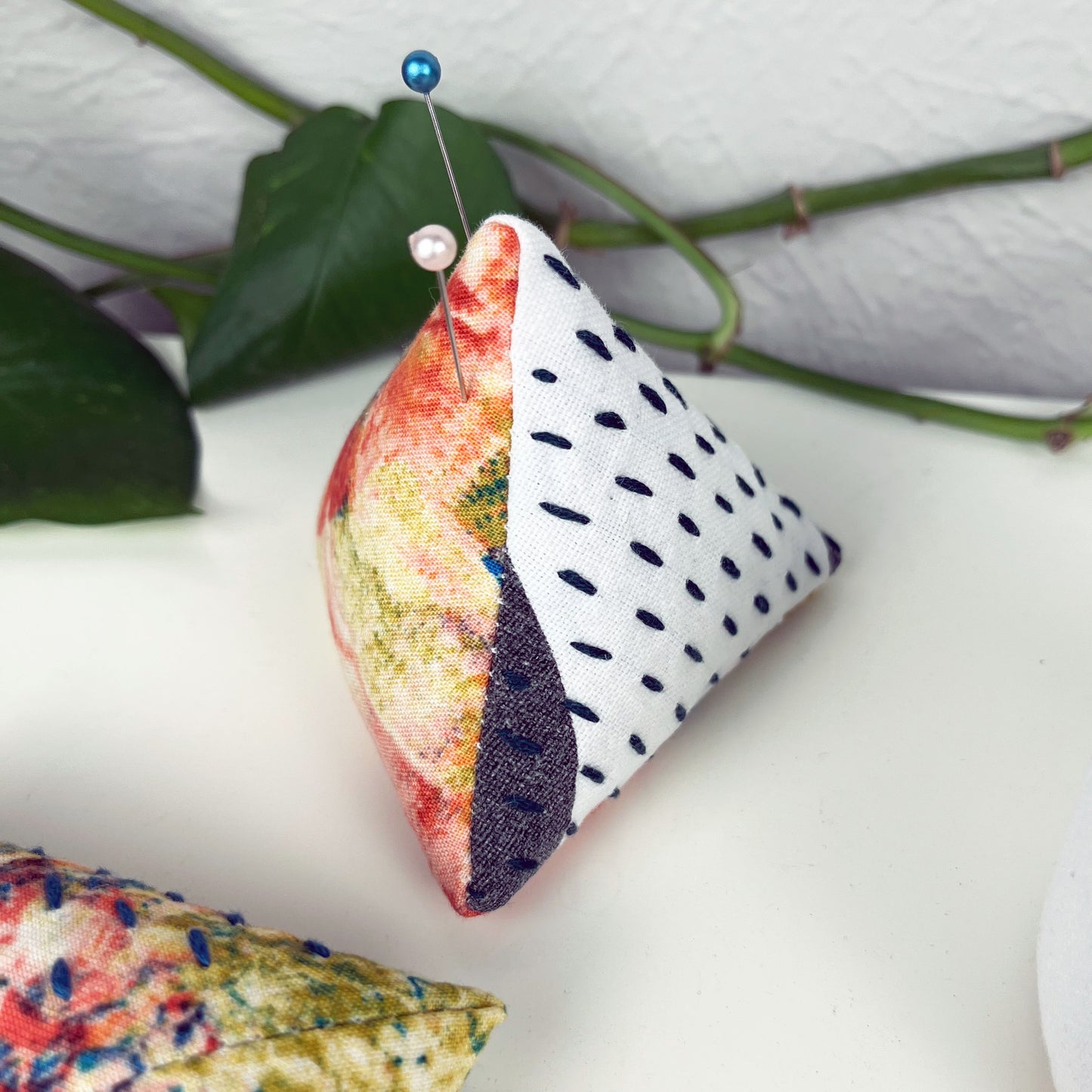 a pyramid pincushion made from multi colored and black and white fabric with contrasting sashiko stitches on a white counter 