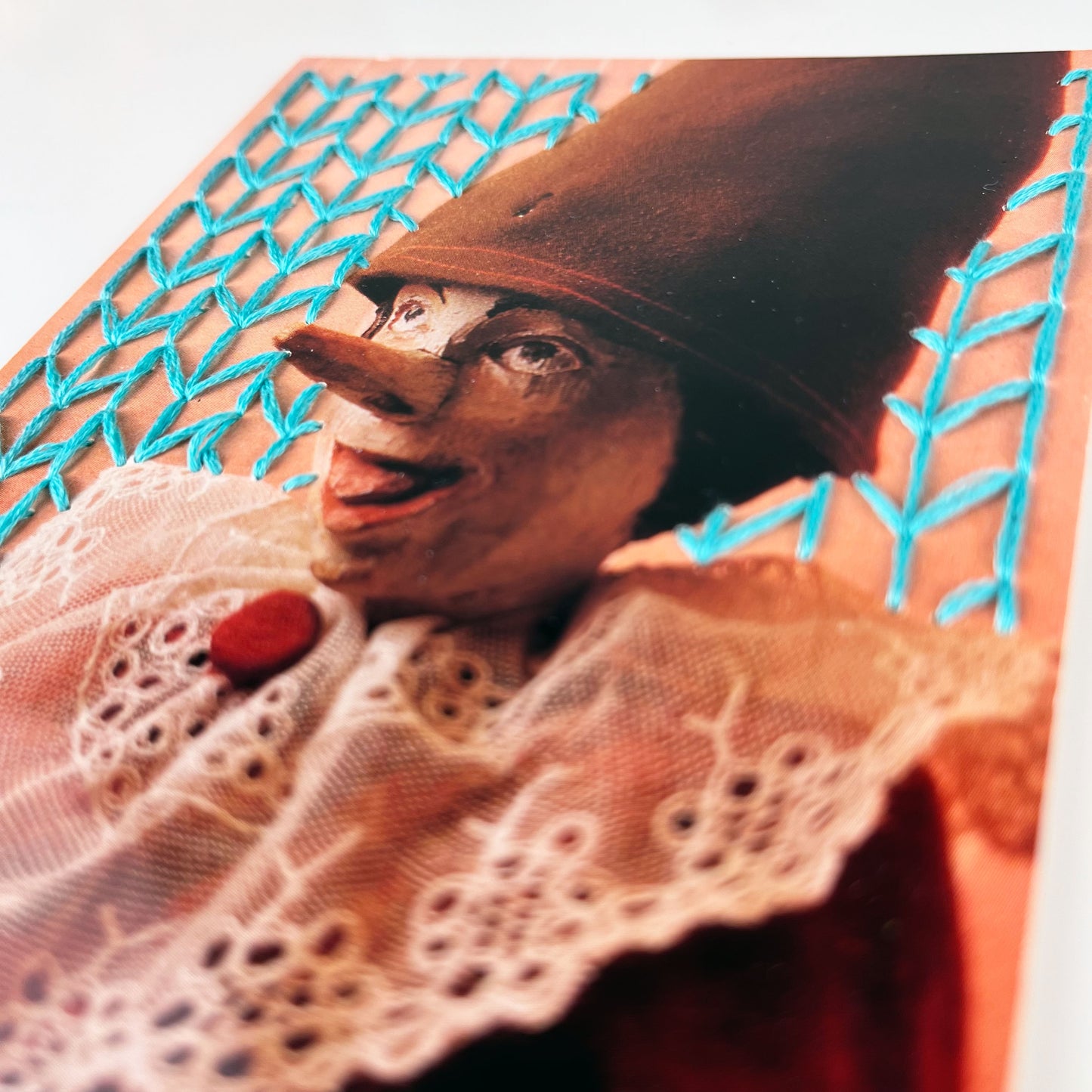 close up angled view of postcard of an old Pinocchio puppet, with the background hand embroidered in aqua in an all over chevron diamond pattern, on a white background
