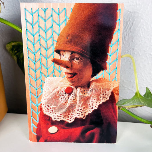 postcard of an old Pinocchio puppet, with the background hand embroidered in aqua in an all over chevron diamond pattern, the postcard has a plant vine behind it
