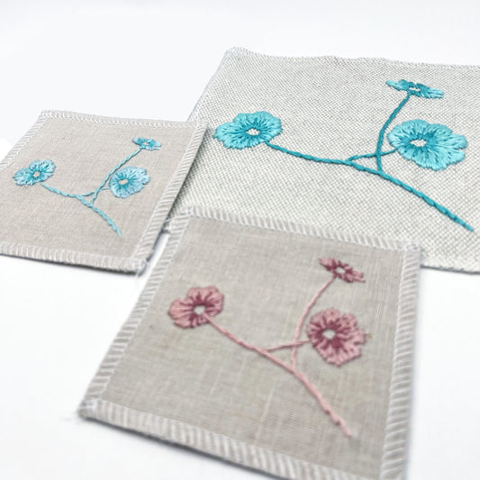 a group of square patches in natural colored fabric, hand embroidered with three poppies on stems, some in shades of pink, some in shades of blue