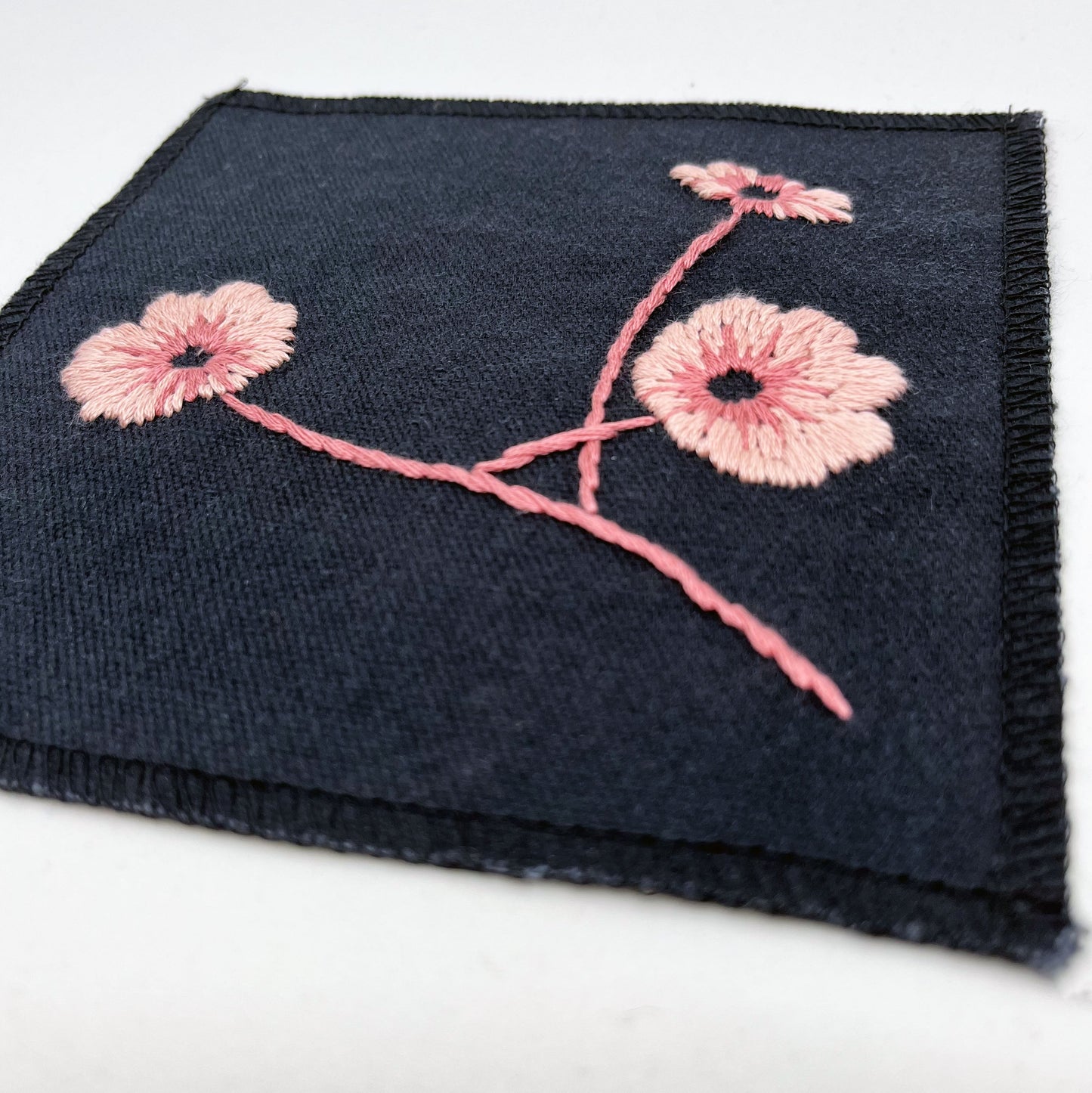 a close up angled view of a black colored square patch, hand embroidered with three poppies on a stem, in shades of pink, with overlocked edges, on a white background