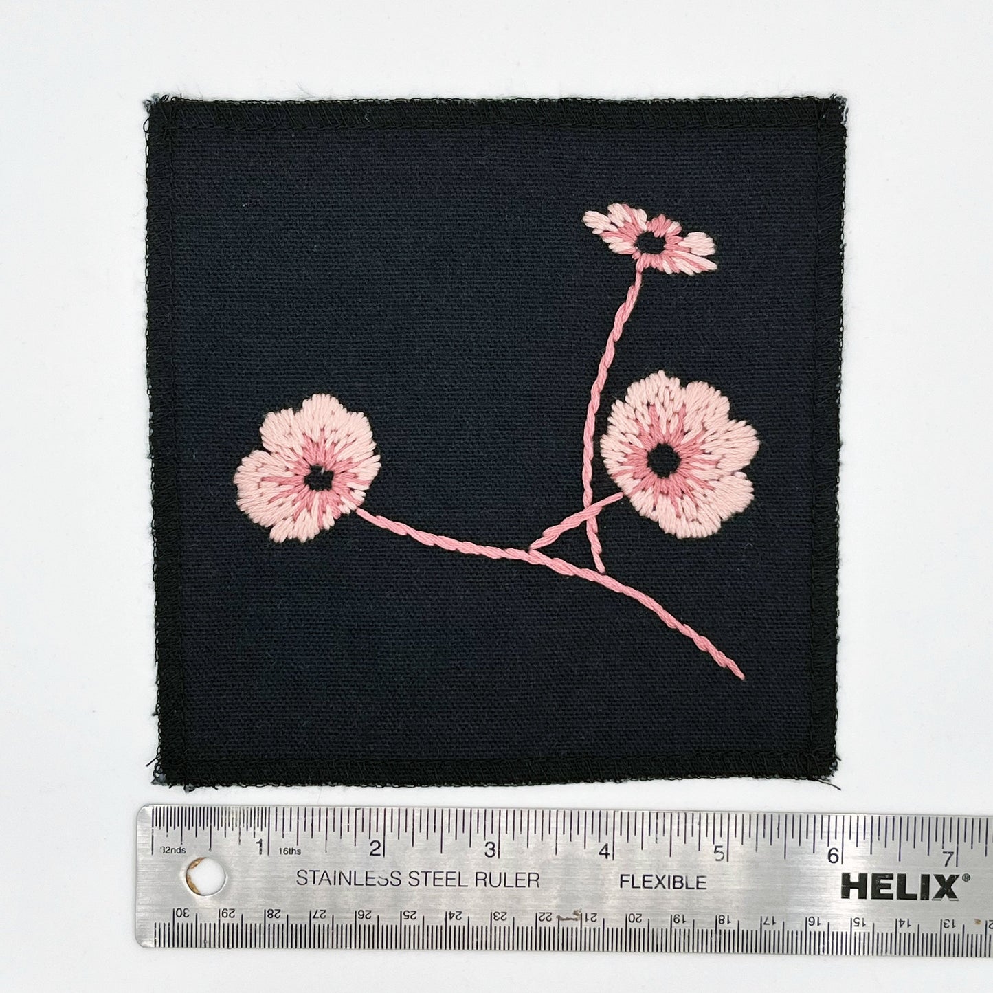 a black square patch, hand embroidered with three poppies on a stem, in shades of pink, with overlocked edges, placed next to a metal ruler to show a width of six inches, on a white background