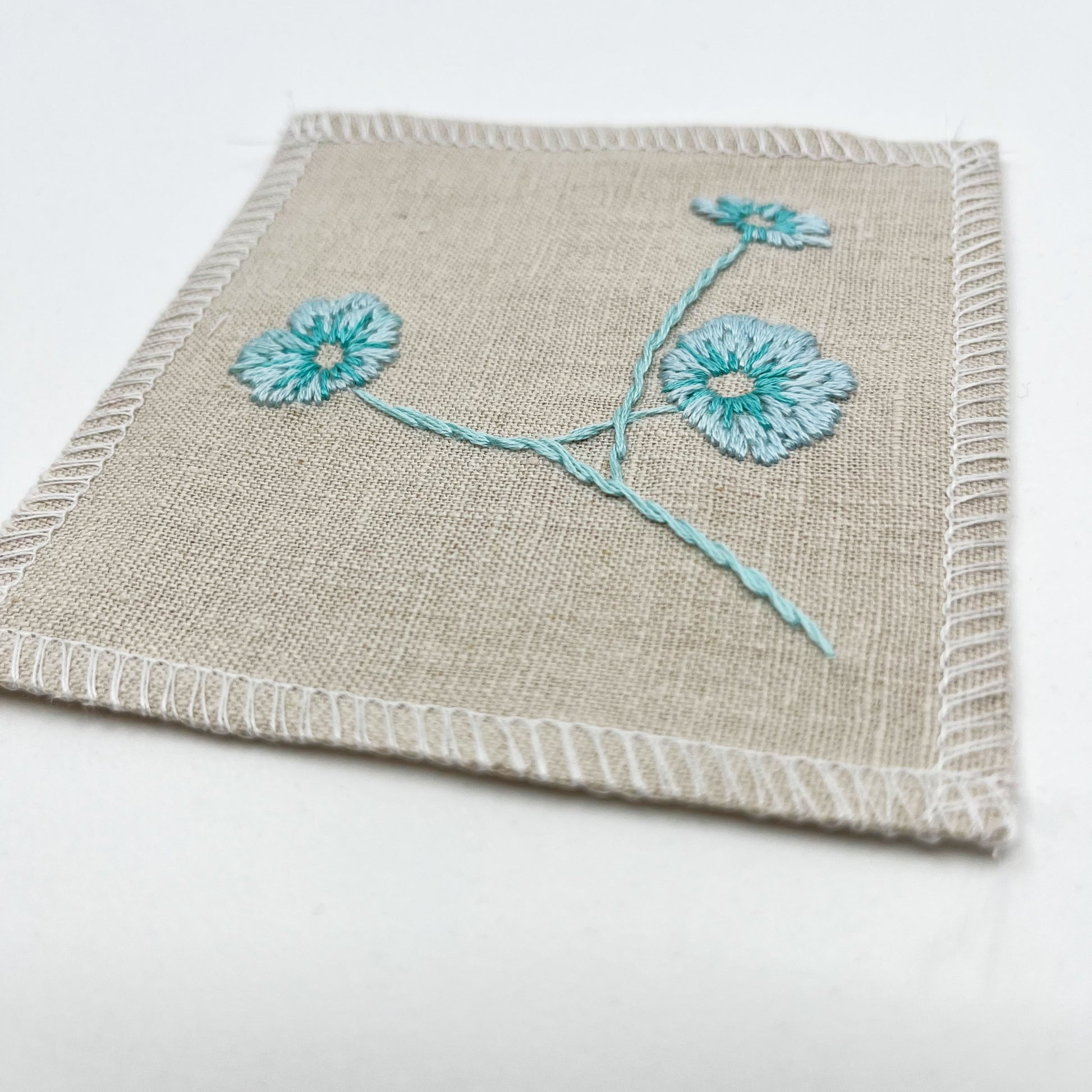 a close up angled view of a natural colored square patch, hand embroidered with three poppies on a stem, in shades of robin's egg blue, with overlocked edges, on a white background