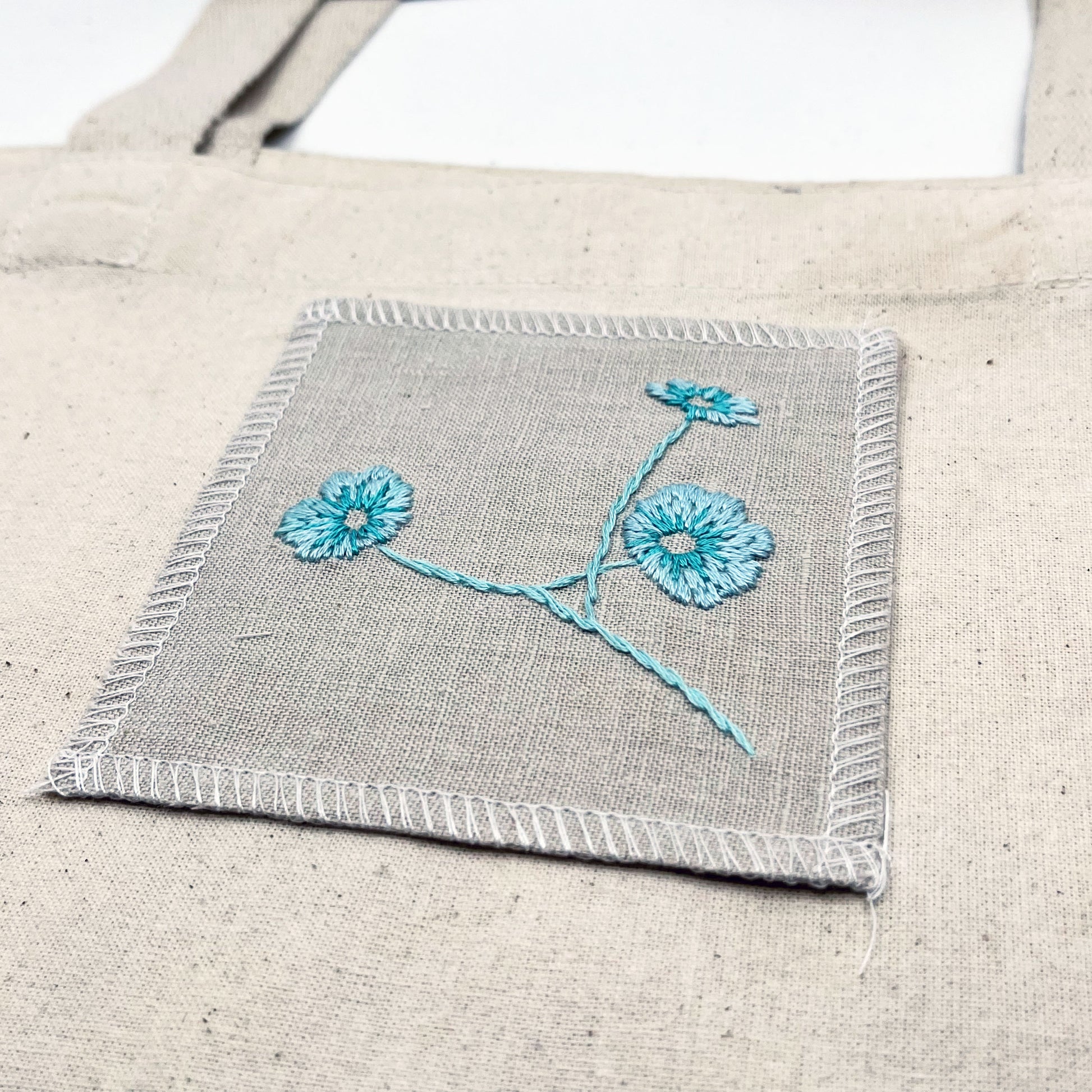 a natural colored square patch, hand embroidered with three poppies on a stem, in shades of robin's egg blue, on a natural canvas tote bag, with overlocked edges