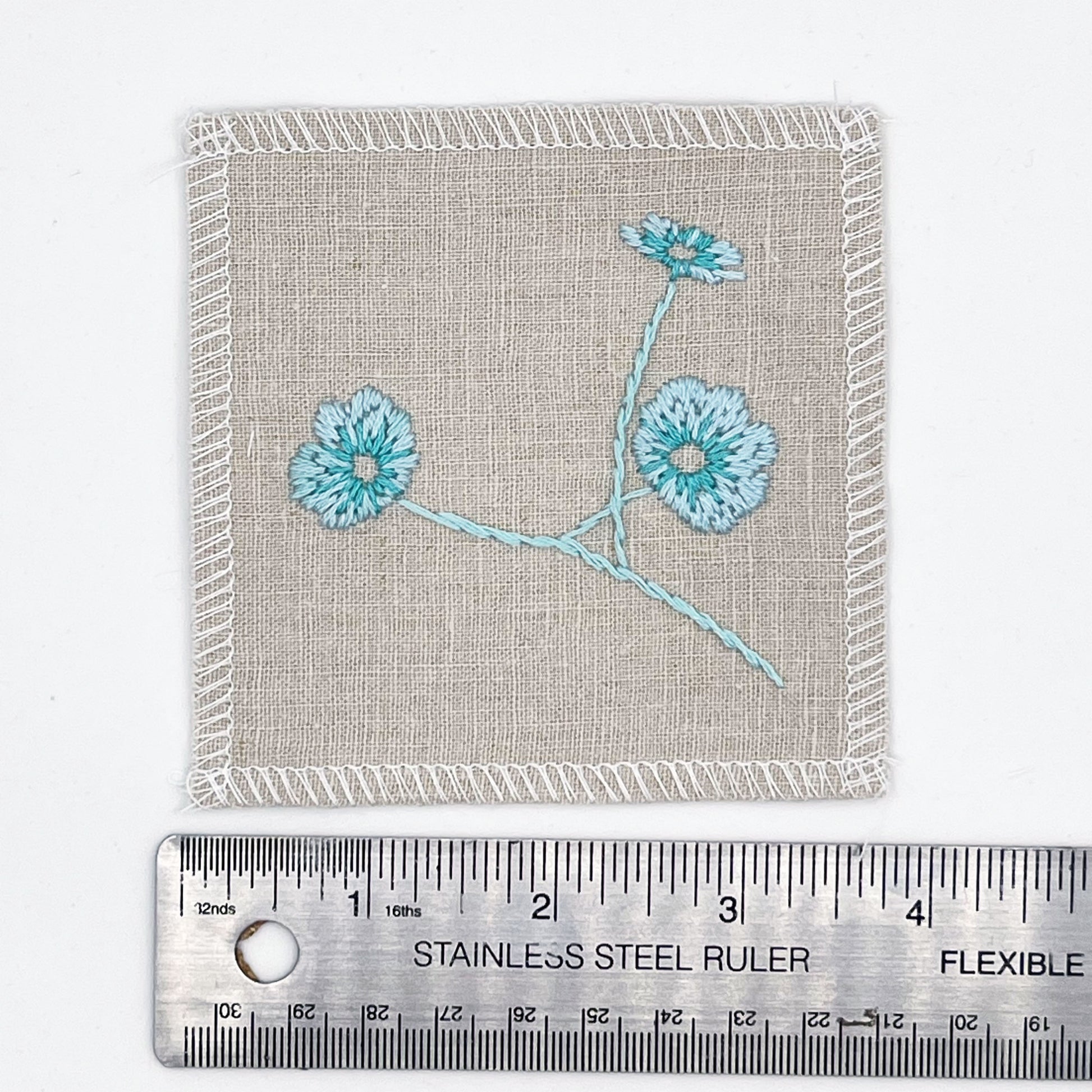 a natural colored square patch, hand embroidered with three poppies on a stem, in shades of robin's egg blue, placed next to a metal ruler to show a width of four inches, on a white background
