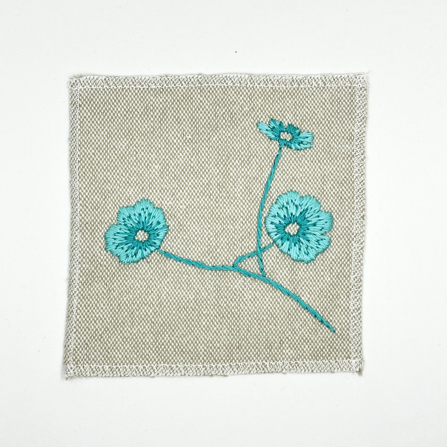 a natural colored square patch, hand embroidered with three poppies on a stem, in shades of robin's egg blue, with overlocked edges, on a white background