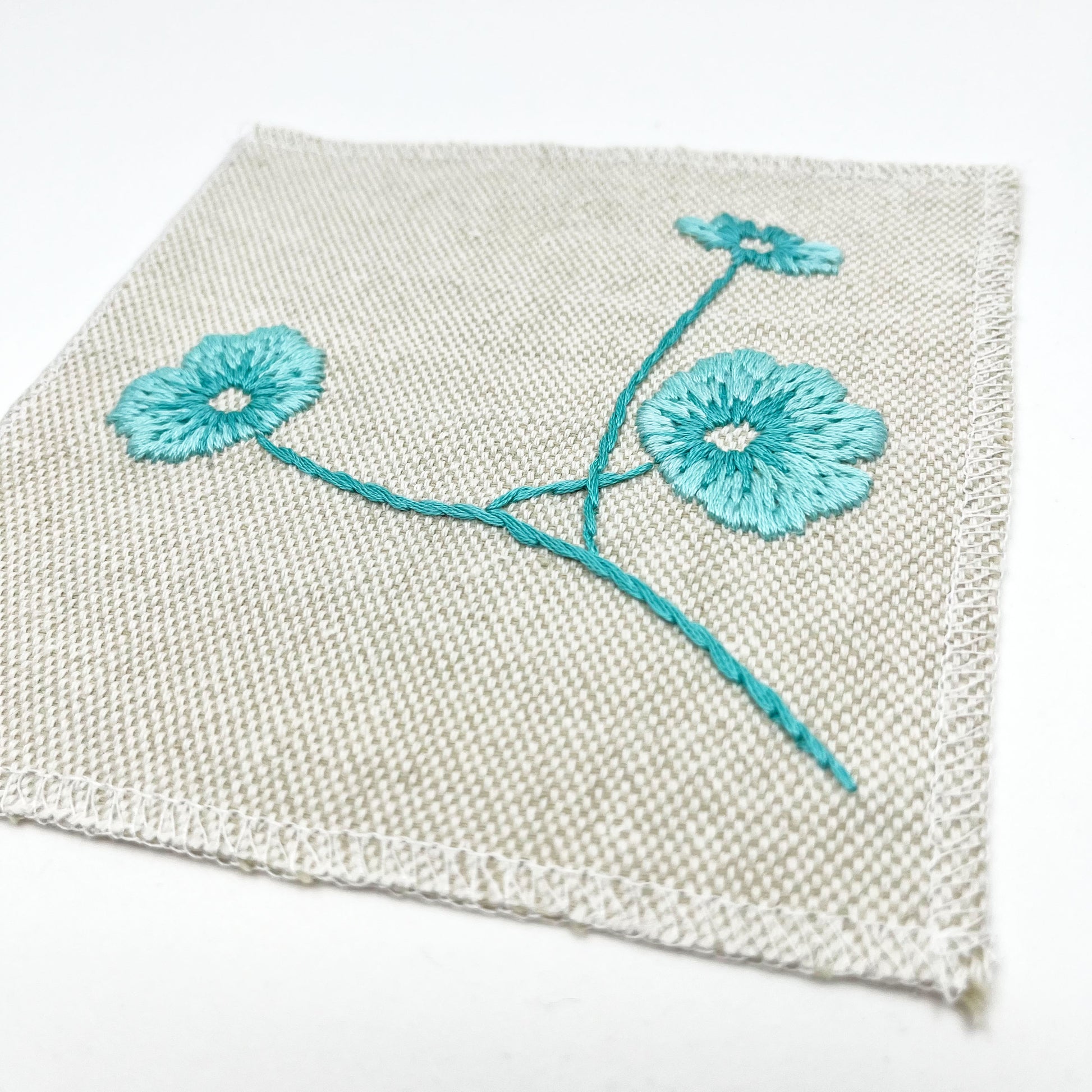 a close up angled view of a natural colored square patch, hand embroidered with three poppies on a stem, in shades of robin's egg blue, with overlocked edges, on a white background