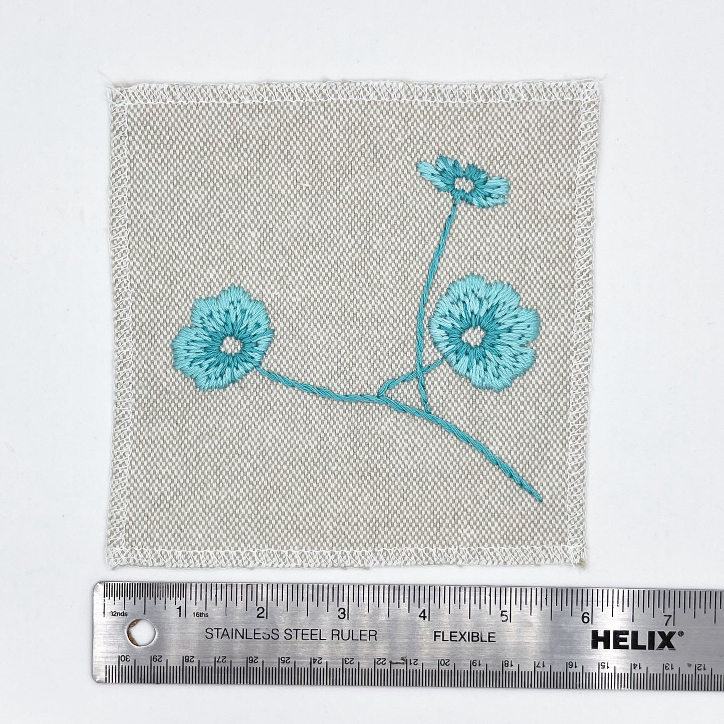a natural colored square patch, hand embroidered with three poppies on a stem, in shades of robin's egg blue, placed next to a metal ruler to show a width of six inches, on a white background