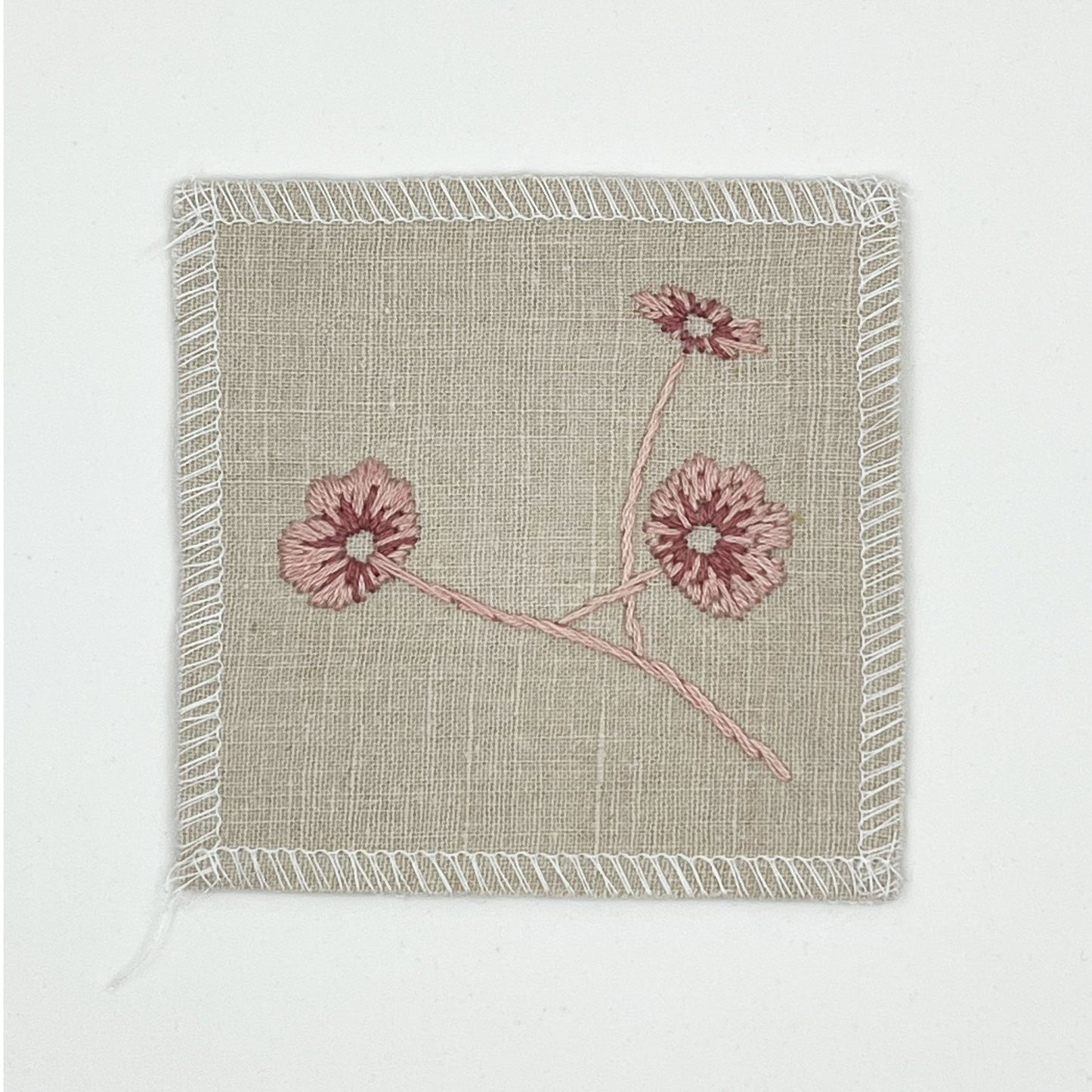 a natural colored square patch, hand embroidered with three poppies on a stem, in shades of pink, with overlocked edges, on a white background