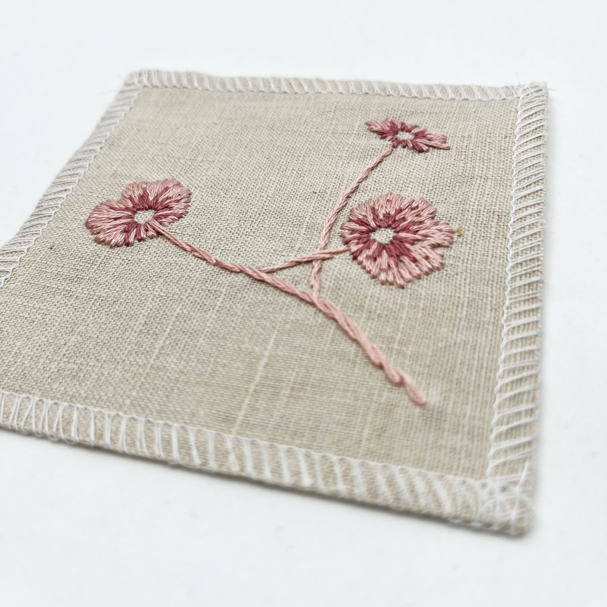 a close up angled view of a natural colored square patch, hand embroidered with three poppies on a stem, in shades of pink, with overlocked edges, on a white background