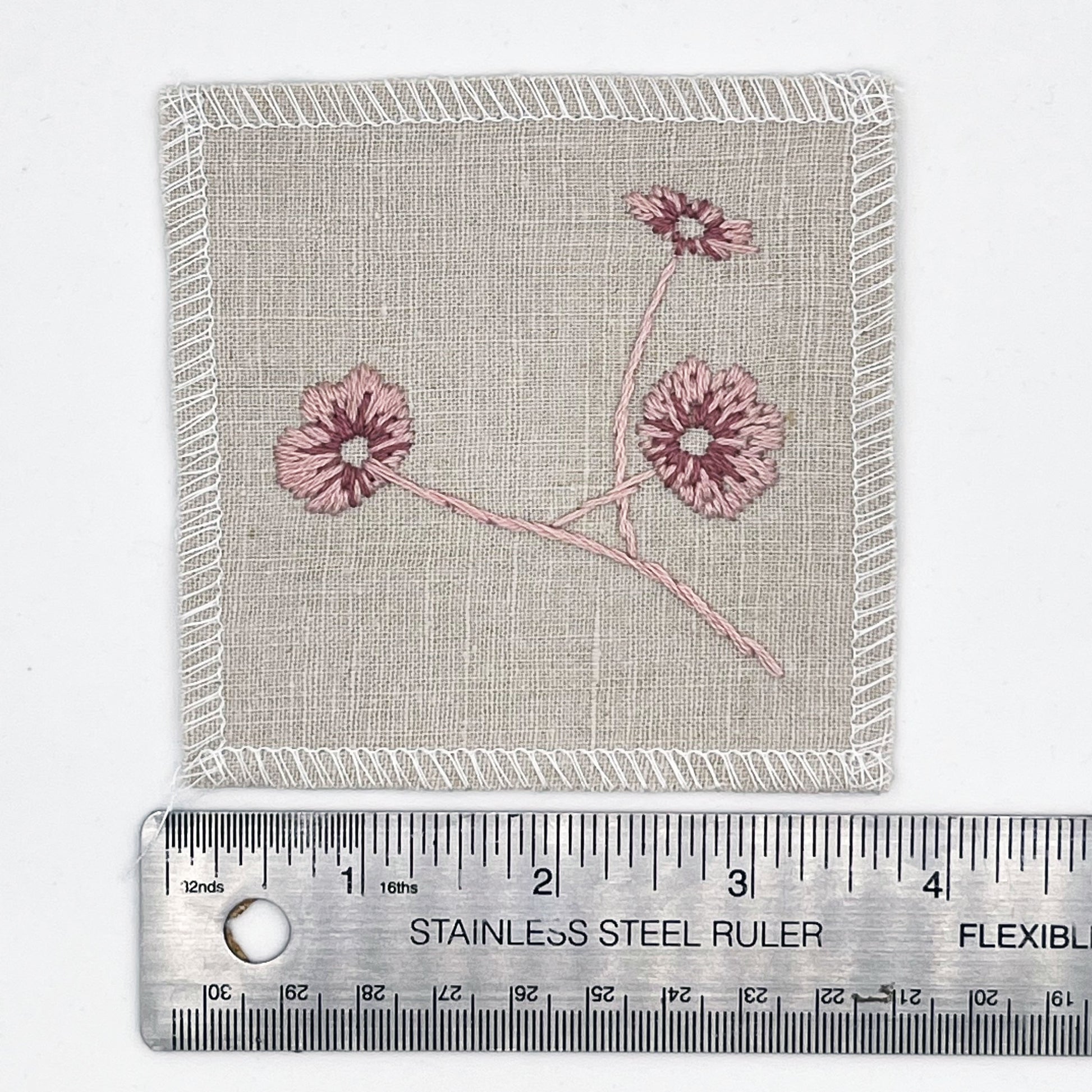 a natural colored square patch, hand embroidered with three poppies on a stem, in shades of pink, with overlocked edges, placed next to a metal ruler to show a width of four inches, on a white background