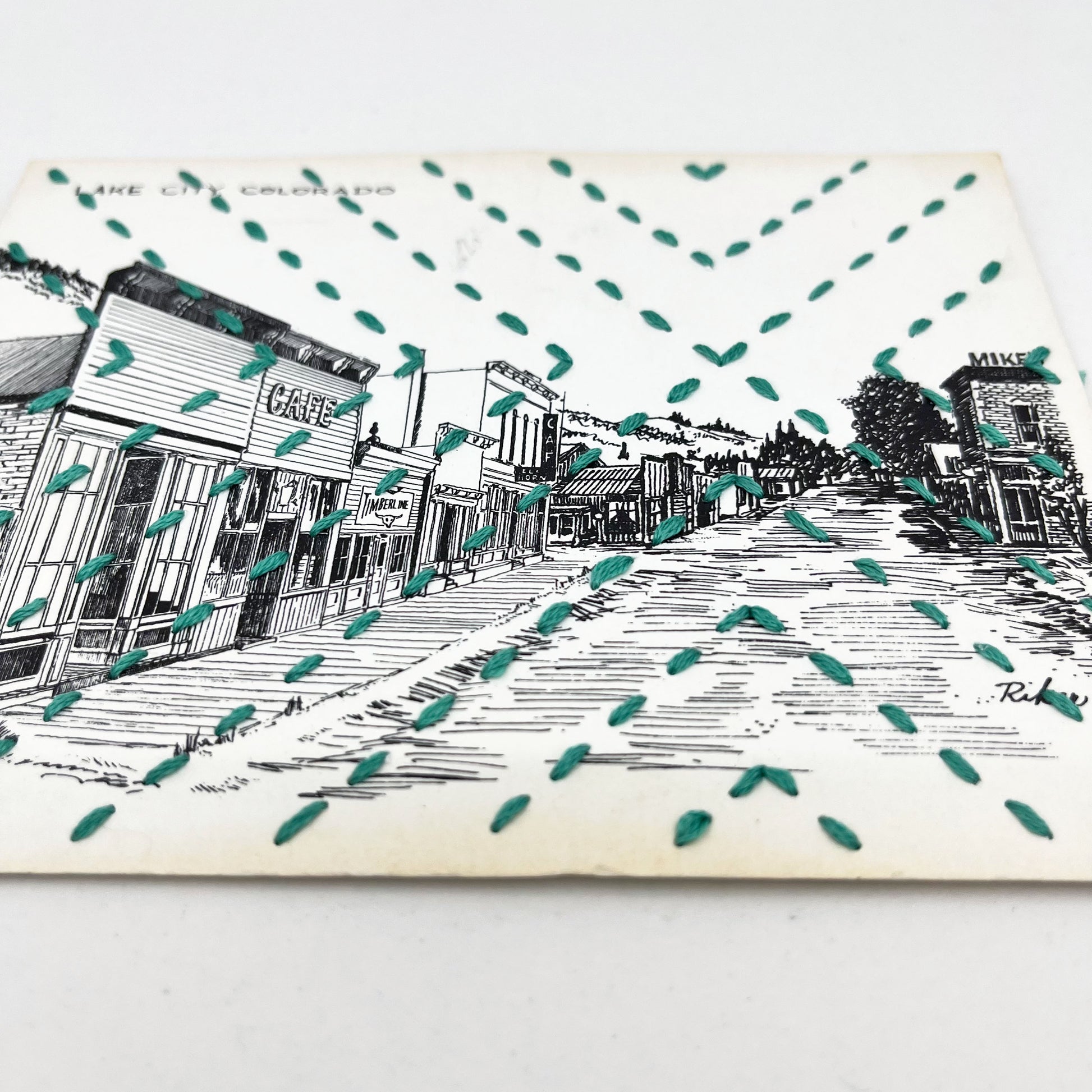 a vintage postcard with a line drawing of Lake City Colorado, hand stitched with a radiating X in green floss