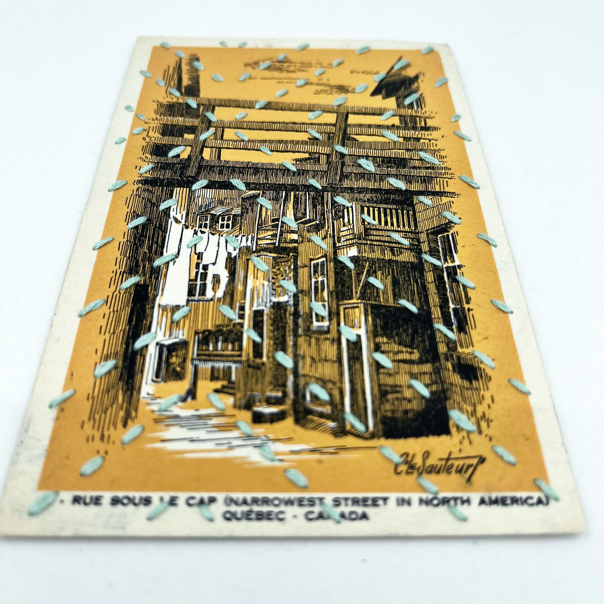 a golden yellow vintage postcard depicting a drawing of an alley in Quebec, stitched over with running stitches in light blue floss in a radiating X pattern