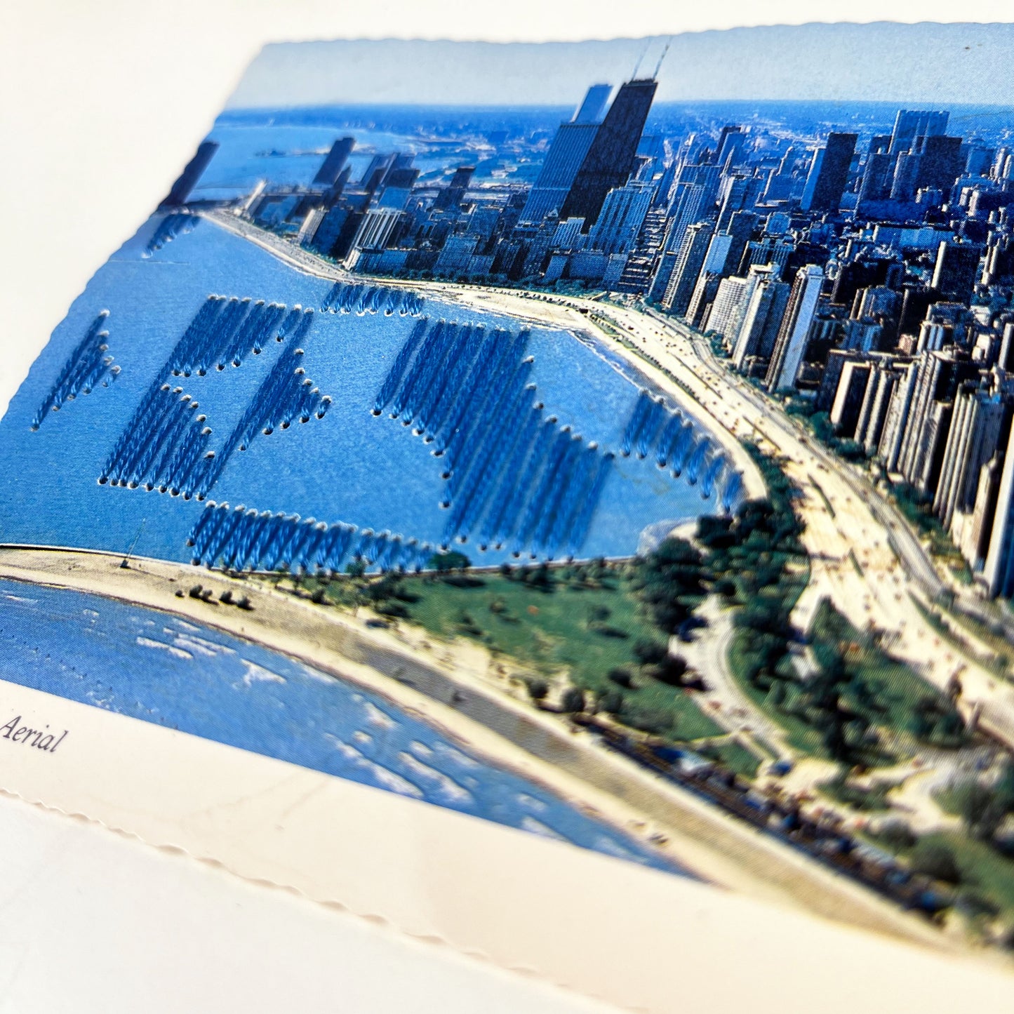 close up angled view of an old postcard of the Chicago skyline along Lake Michigan, with part of the lake hand embroidered over with an abstract partial circle design in blue