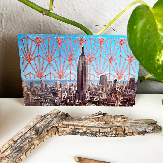 an old postcard of the Empire State Building, with a hand embroidered Art Deco design in coral stitched in the background on the sky only, the postcard is sitting on a white dresser with a plant vine behind it and sticks and dried flowers in front of it