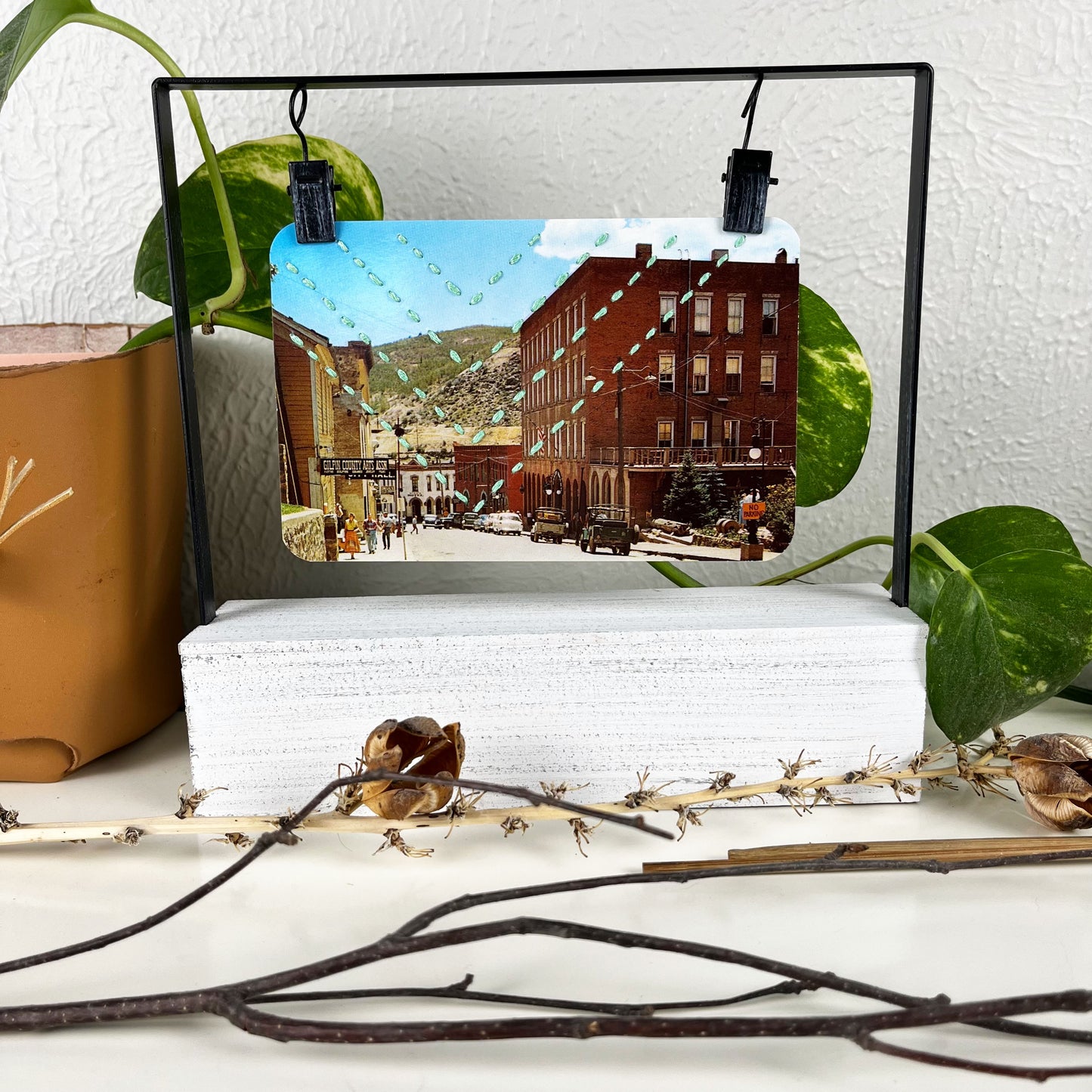 a vintage postcard of a small mountain town in Colorado, hand stitched with four rows of chevrons in aqua floss, in a clip picture frame, on a white counter surrounded by dried sticks and yucca, with a pothos vine behind it