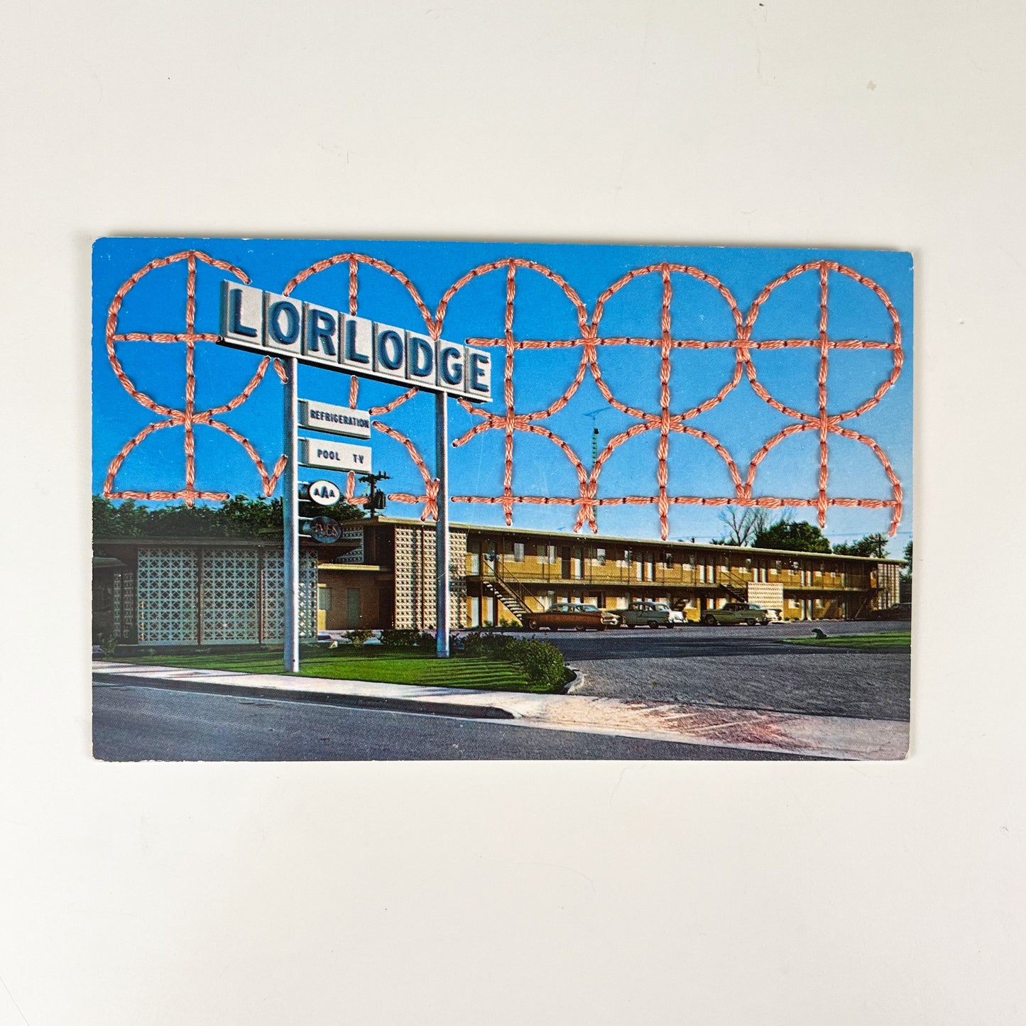 an old postcard of a motel called Lorlodge, with a hand embroidered circle and line design like breeze blocks in peach stitched in the background on the sky only, the postcard is sitting on a white background