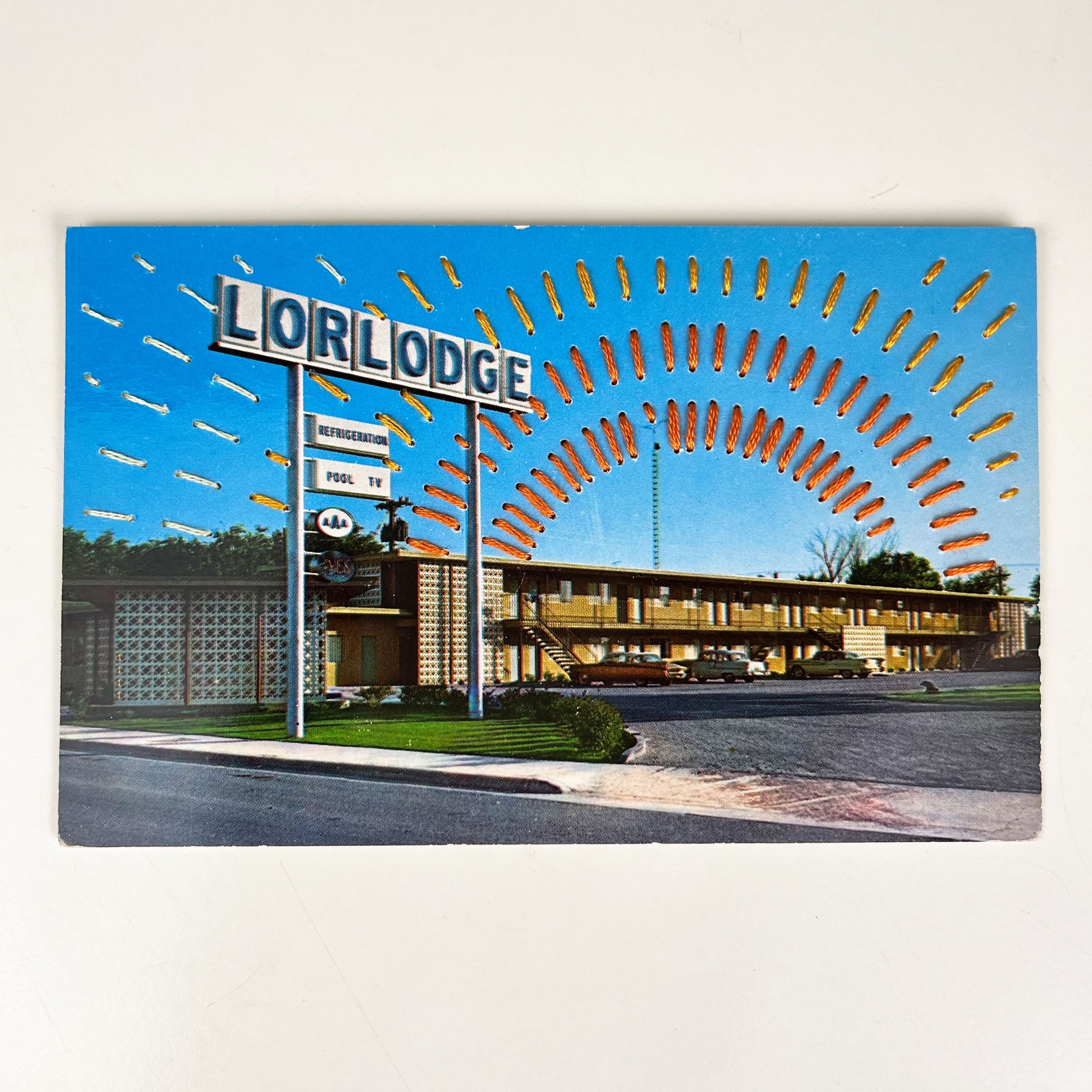 an old postcard of a motel called Lorlodge, with hand embroidered dashed lined resembling sun beams in oranges and yellows, stitched in the background on the sky only, the postcard is sitting on a white background