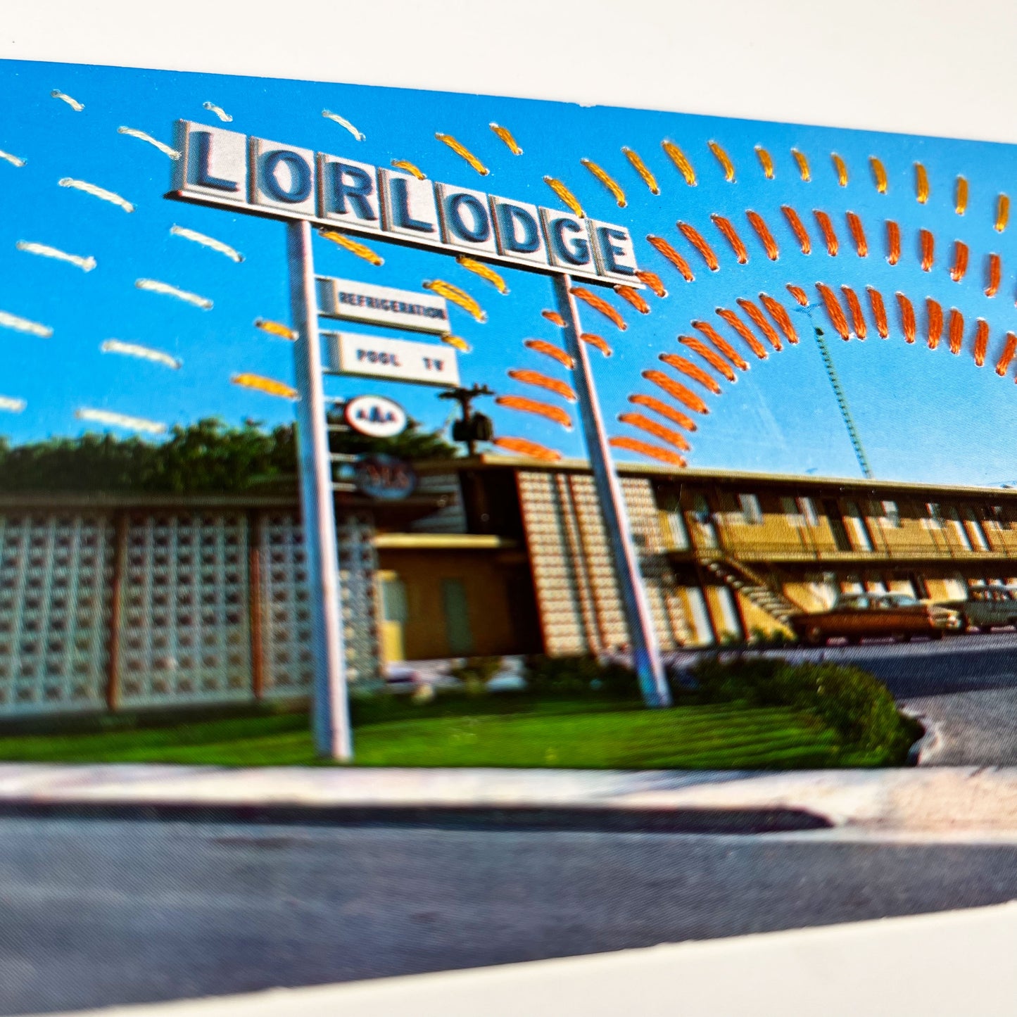 close up angled view of an old postcard of a motel called Lorlodge, with hand embroidered dashed lined resembling sun beams in oranges and yellows, stitched in the background on the sky only, the postcard is sitting on a white background