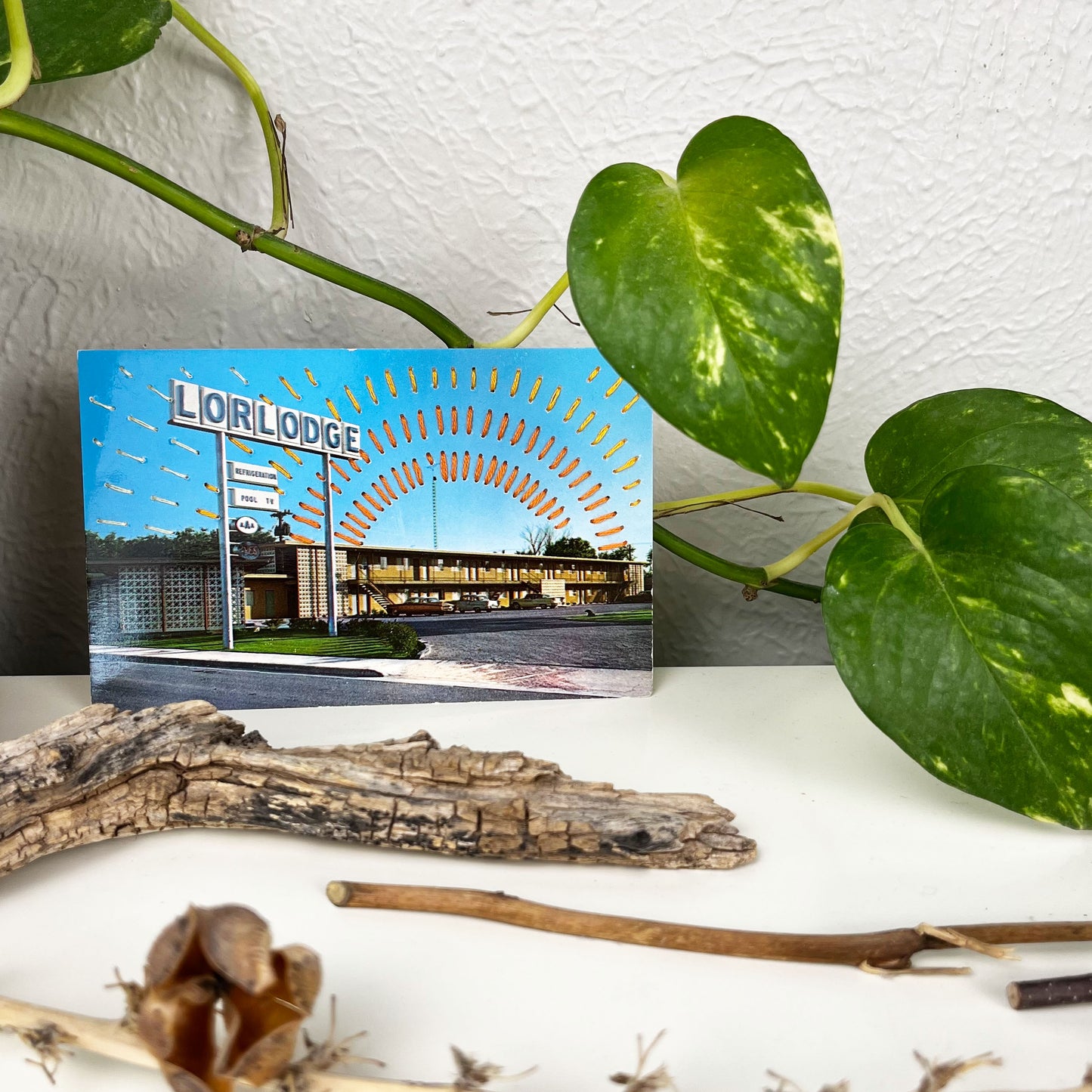 an old postcard of a motel called Lorlodge, with hand embroidered dashed lined resembling sun beams in oranges and yellows, stitched in the background on the sky only, the postcard is sitting on a white dresser with a plant vine behind it and sticks and dried flowers in front of it