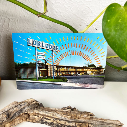 an old postcard of a motel called Lorlodge, with hand embroidered dashed lined resembling sun beams in oranges and yellows, stitched in the background on the sky only, the postcard is sitting on a white dresser with a plant vine behind it and sticks and dried flowers in front of it