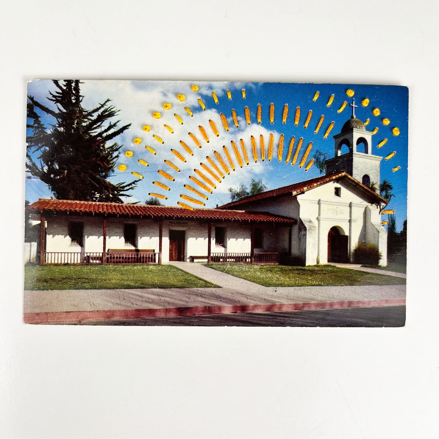 an old postcard of a mission building in Santa Cruz, with hand embroidered dashed lines and french knots resembling sun beams in oranges and yellows, stitched in the background on the sky only, the postcard is sitting on a white background