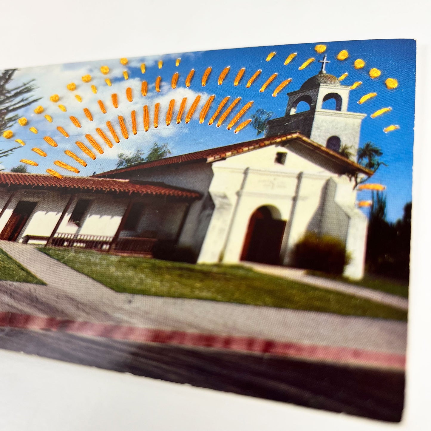 close up angled view of an old postcard of a mission building in Santa Cruz, with hand embroidered dashed lines and french knots resembling sun beams in oranges and yellows, stitched in the background on the sky only, the postcard is sitting on a white background