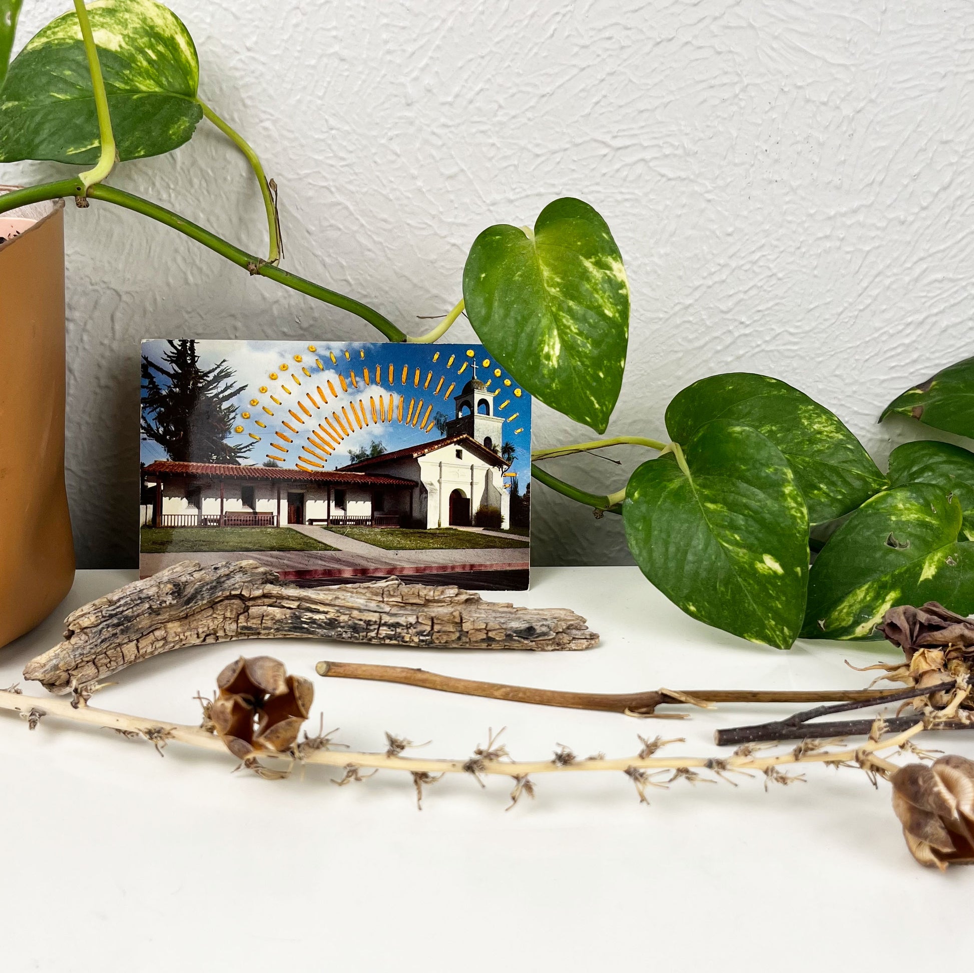 an old postcard of a mission building in Santa Cruz, with hand embroidered dashed lines and french knots resembling sun beams in oranges and yellows, stitched in the background on the sky only, the postcard is sitting on a white dresser with a plant vine behind it and dried flowers and sticks in front of it