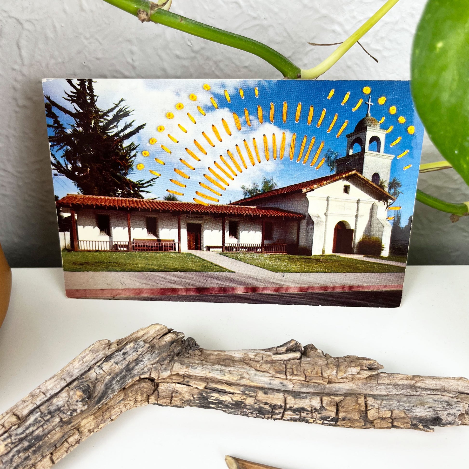 an old postcard of a mission building in Santa Cruz, with hand embroidered dashed lines and french knots resembling sun beams in oranges and yellows, stitched in the background on the sky only, the postcard is sitting on a white dresser with a plant vine behind it and dried flowers and sticks in front of it