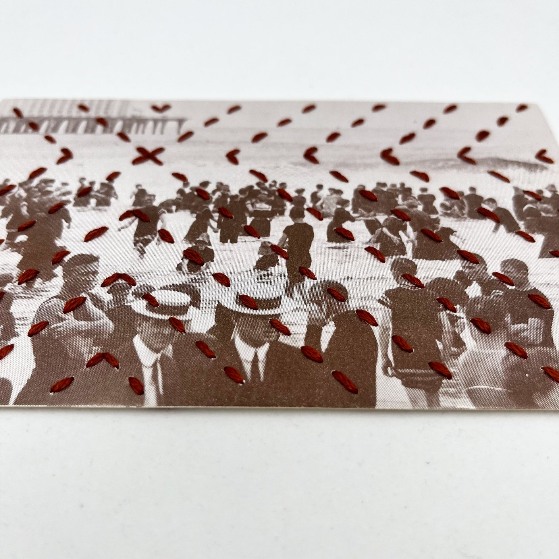 a vintage postcard of a crowd of people in the water in Atlantic City, hand stitched over with a radiating X pattern in brick red