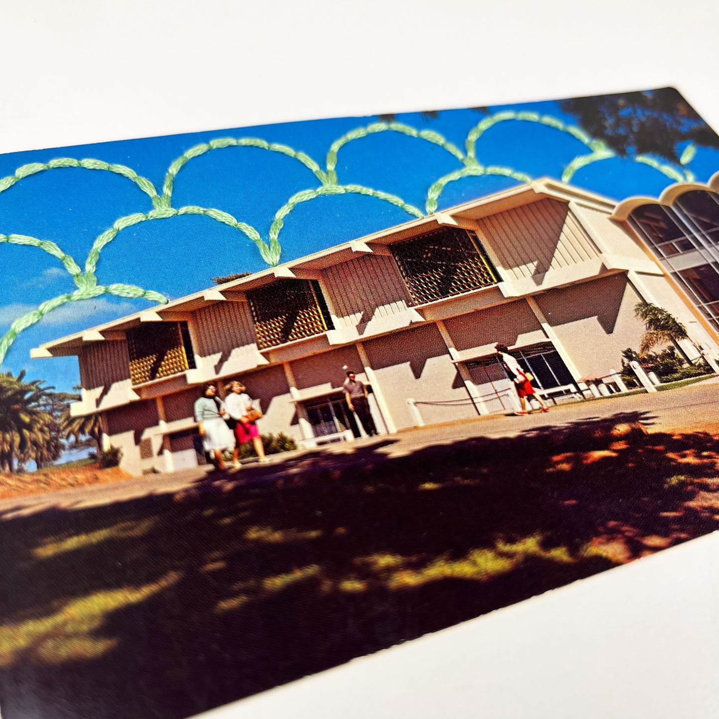 close up angled view of an old postcard of a building at a college in San Diego, with a hand embroidered overlapping circle design like scales in mint green stitched in the background on the sky only, the postcard is sitting on a white background