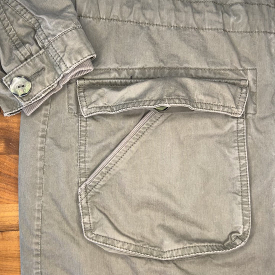 view of a pocket on an olive colored jacket, hands appear holding a square natural colored patch with embroidered triangles, turn the patch to show the back, and then place it face up on the pocket