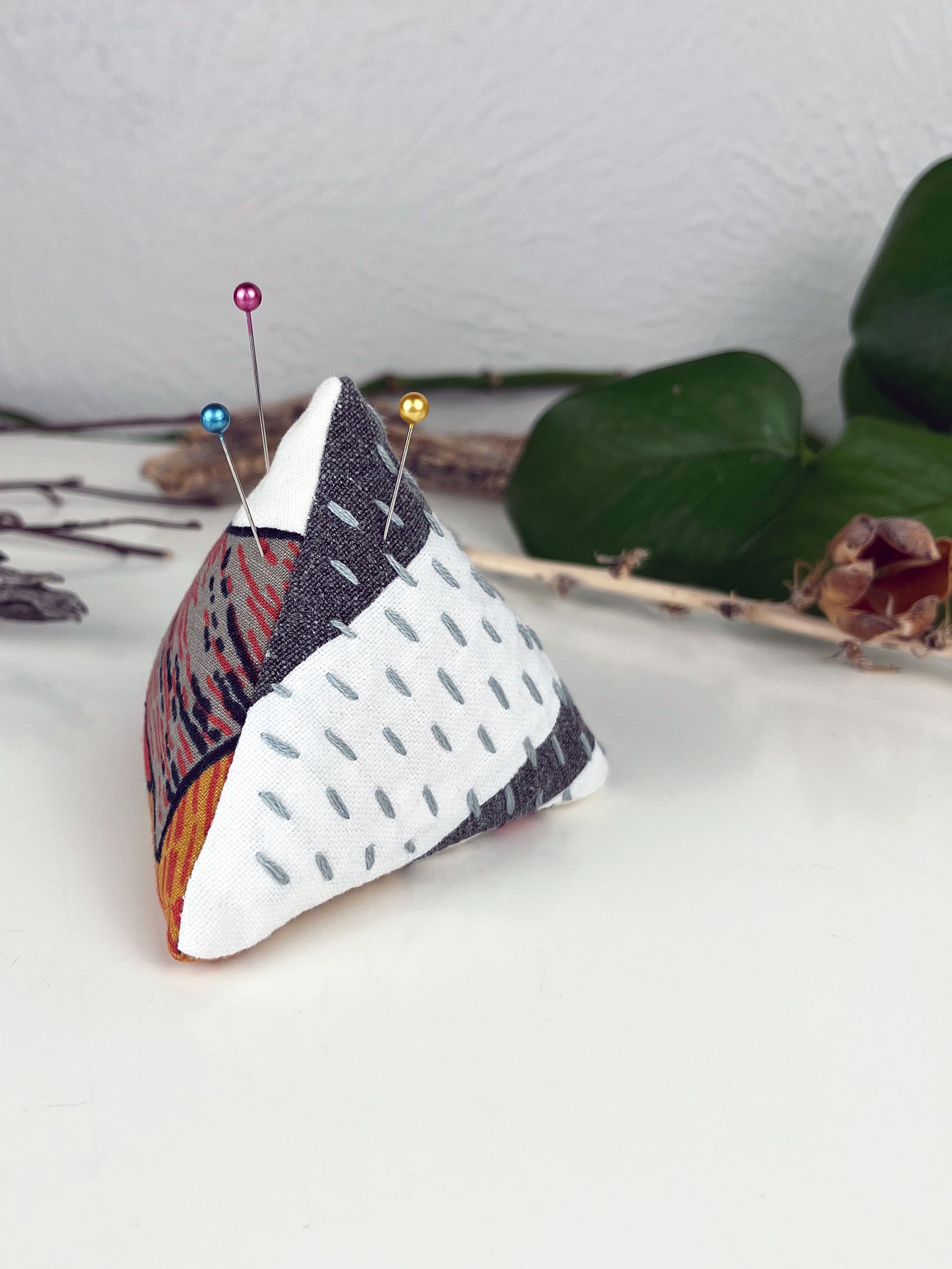 a pyramid pincushion made from floral and black and white fabric with contrasting sashiko stitches on a white counter with dried twigs seedpods and a pothos plant in the background