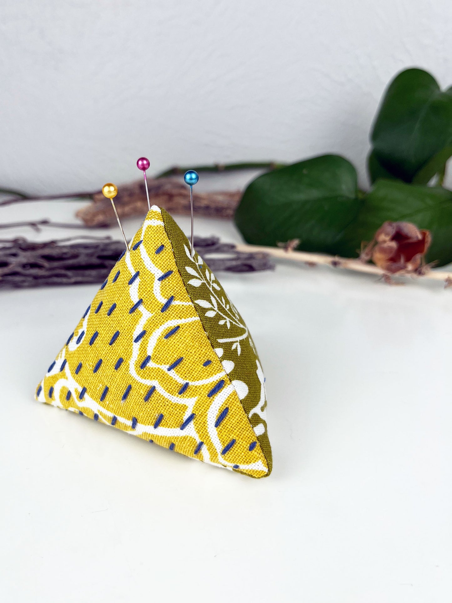a pyramid pincushion made from floral olive and mustard fabric with contrasting sashiko stitches on a white counter with dried twigs seedpods and a pothos plant in the background